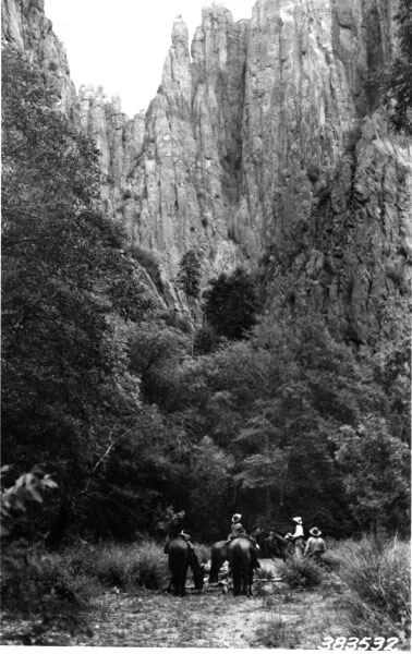 Una fotografía histórica en blanco y negro que muestra a un pequeño grupo de personas montando a caballo por un sendero de tierra al pie de altos y escarpados acantilados rocosos.