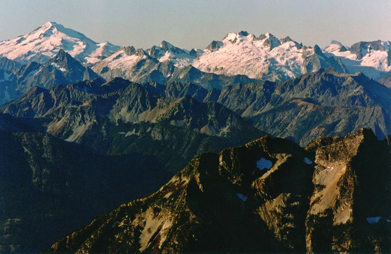 A expansive high-angle view of multiple snow-capped mountain peaks and deep valleys under a clear sky.