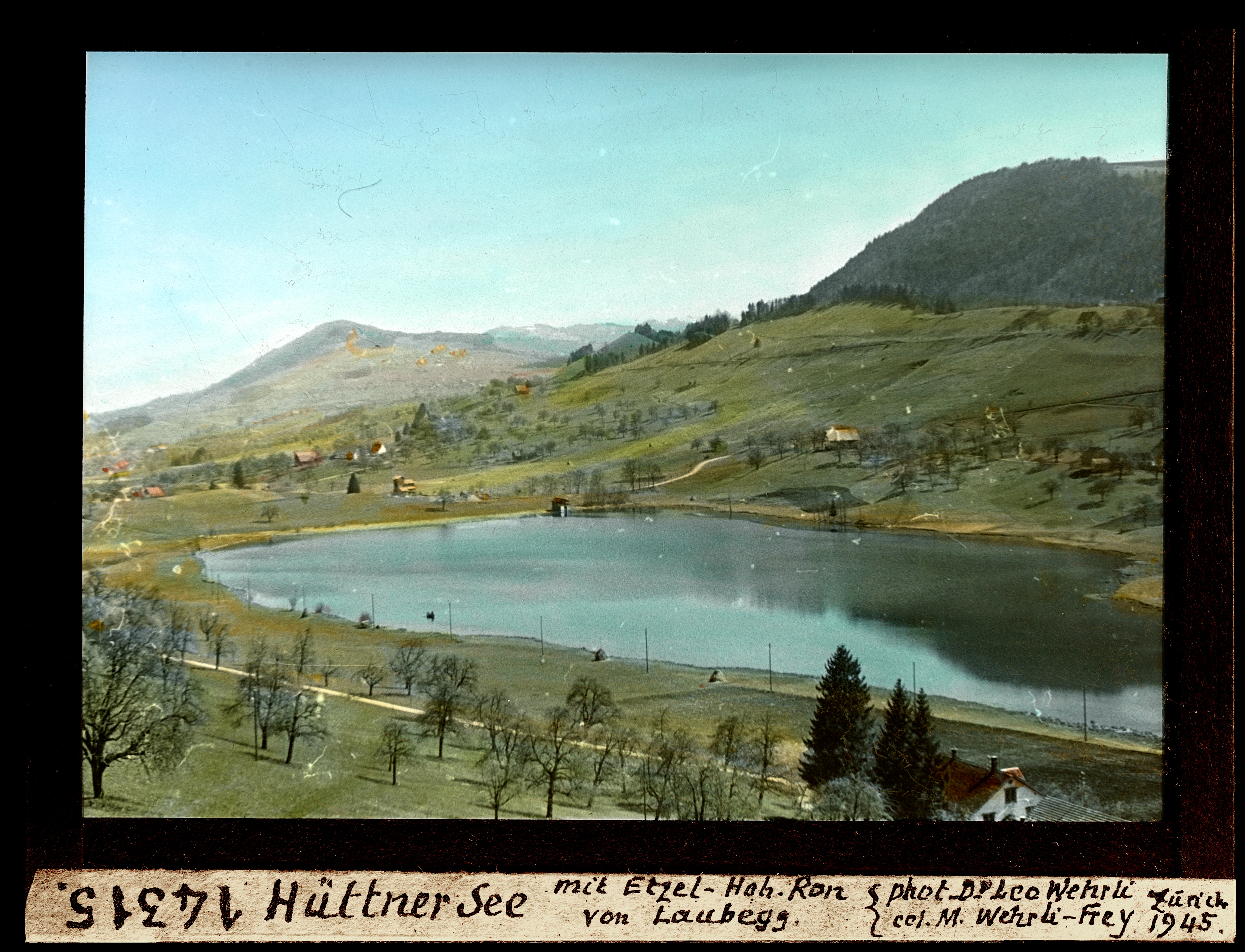 An archival landscape photograph showing a small lake surrounded by rolling green hills, pastures, and trees, with a mountain ridge in the distance.