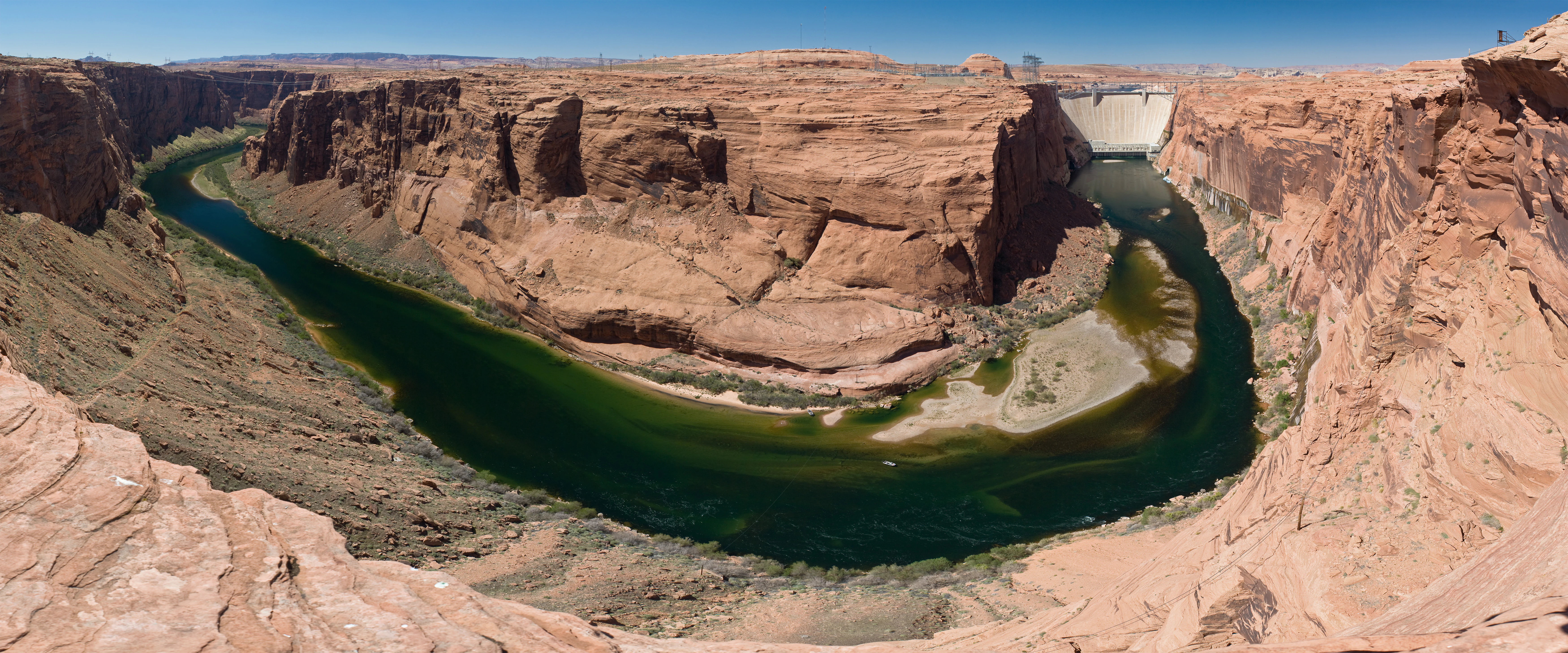 Panoramic view of a deep canyon with a winding green river at the bottom and a large concrete dam visible in the distance.