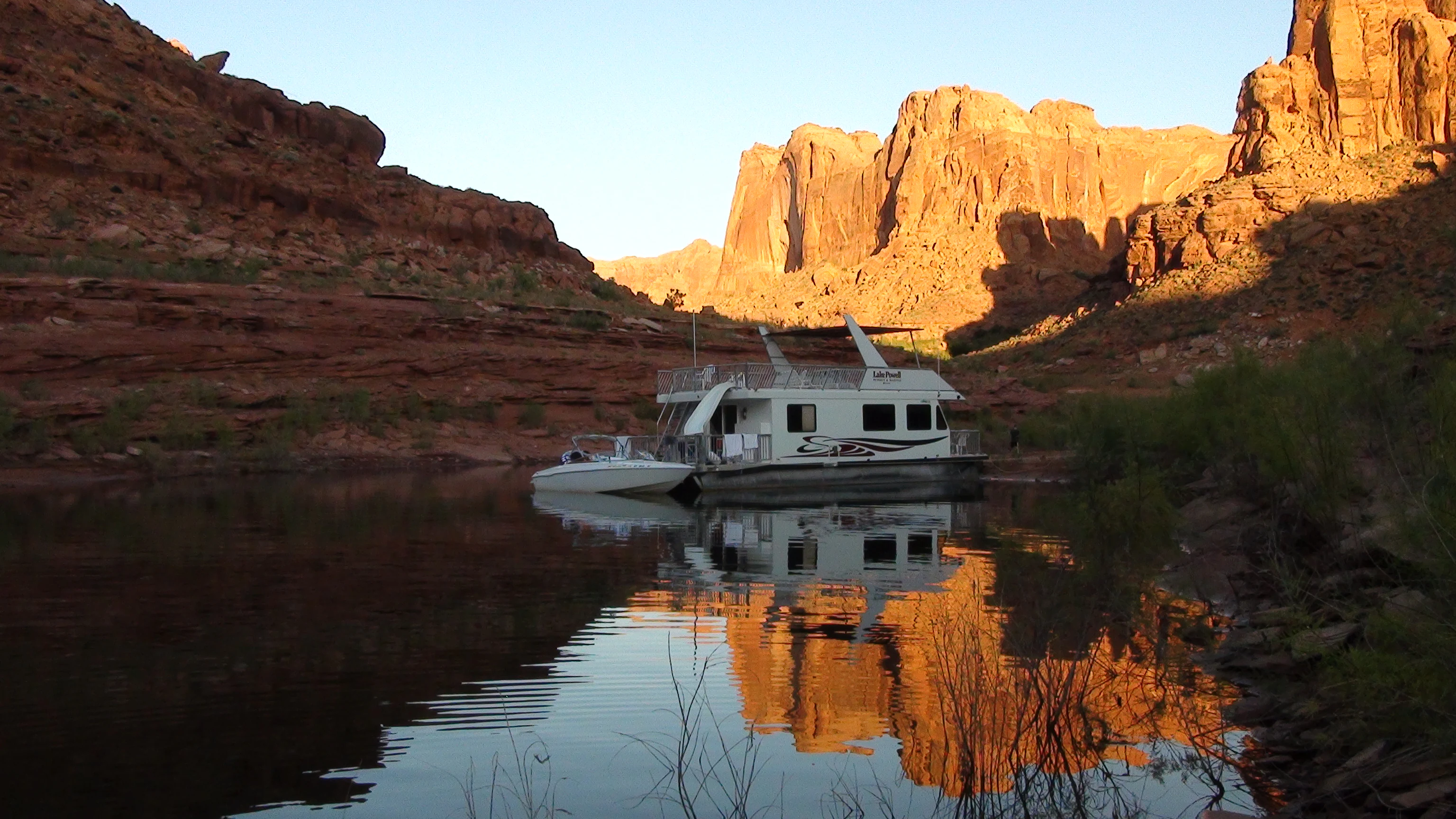 A houseboat and a small motorboat docked in a narrow canyon on a calm lake, with red sandstone cliffs reflecting in the water during golden hour.