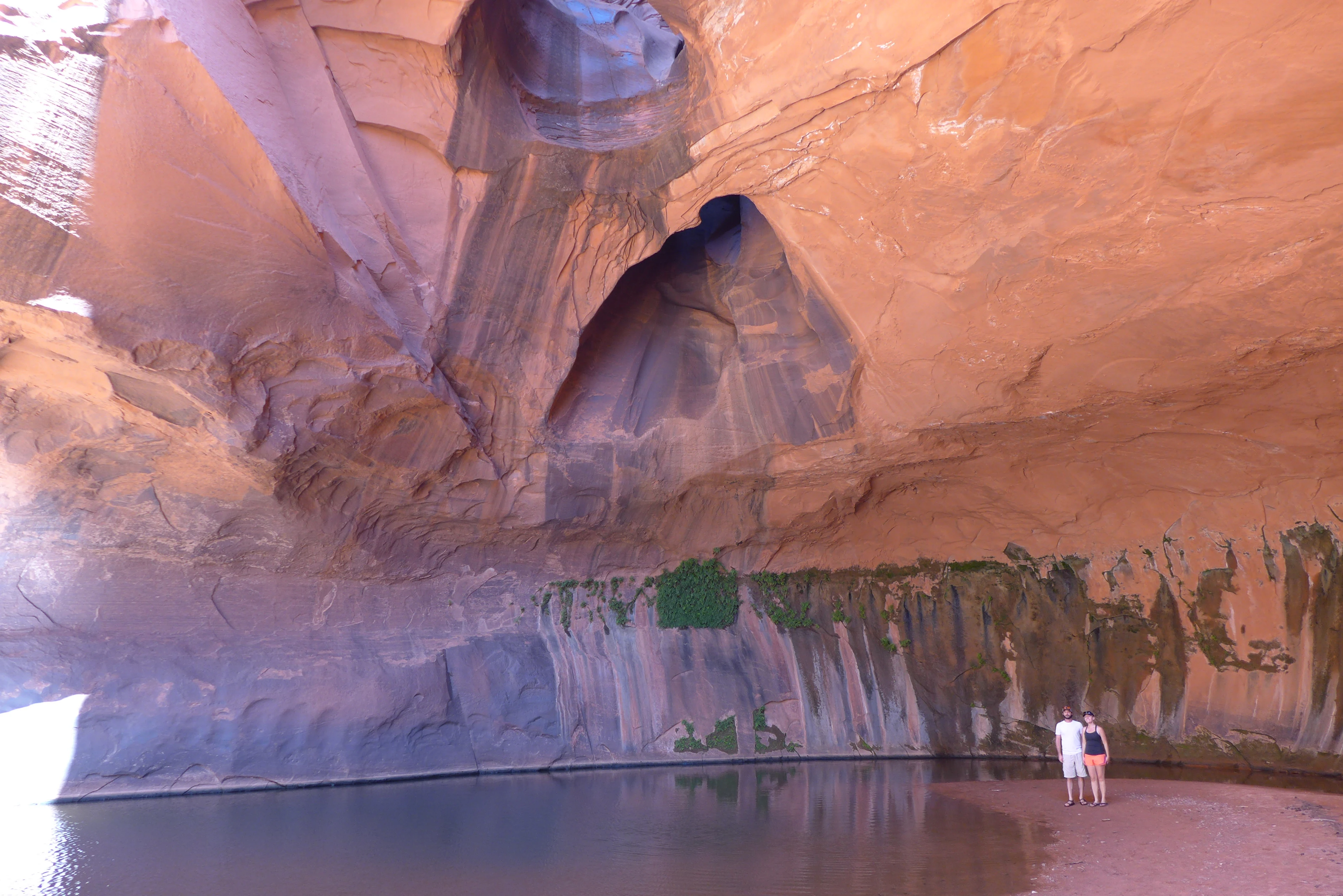 A wide view of the Golden Cathedral, a large orange sandstone alcove with deep pools of water at the base and two distinct circular openings in the high ceiling that allow light to stream into the shaded space. Two hikers stand on the shore near the water.
