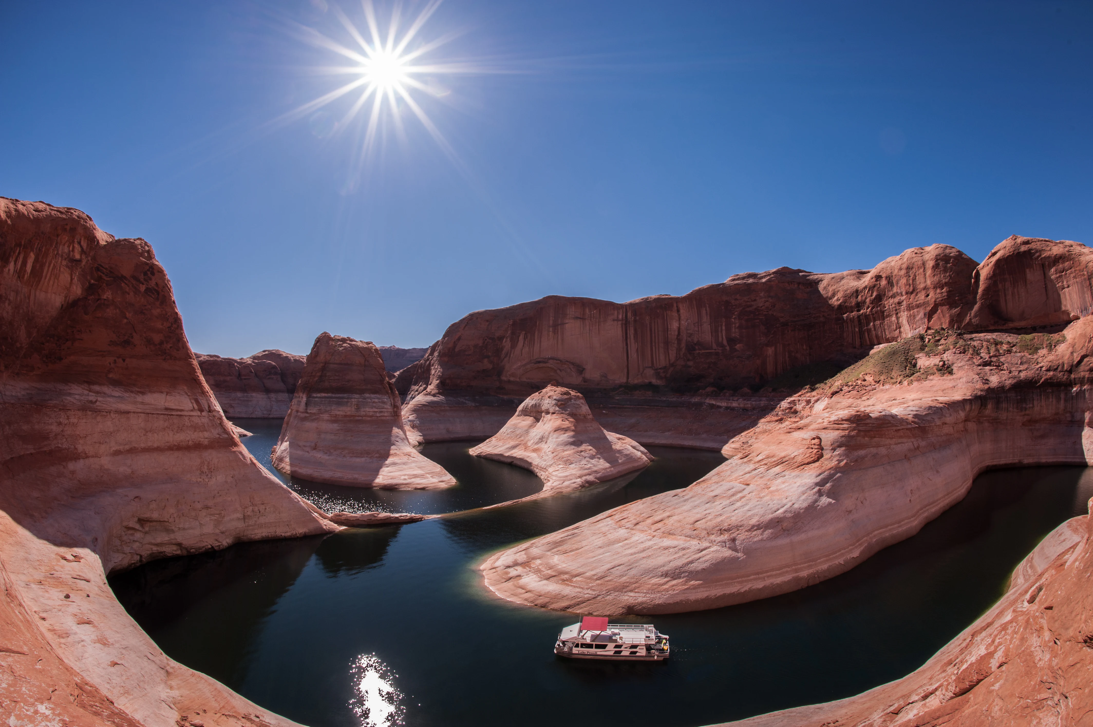 A wide angle shot of a white houseboat in a narrow, winding section of Lake Powell, bordered by deep orange sandstone cliffs under a clear blue sky with a bright sun star.
