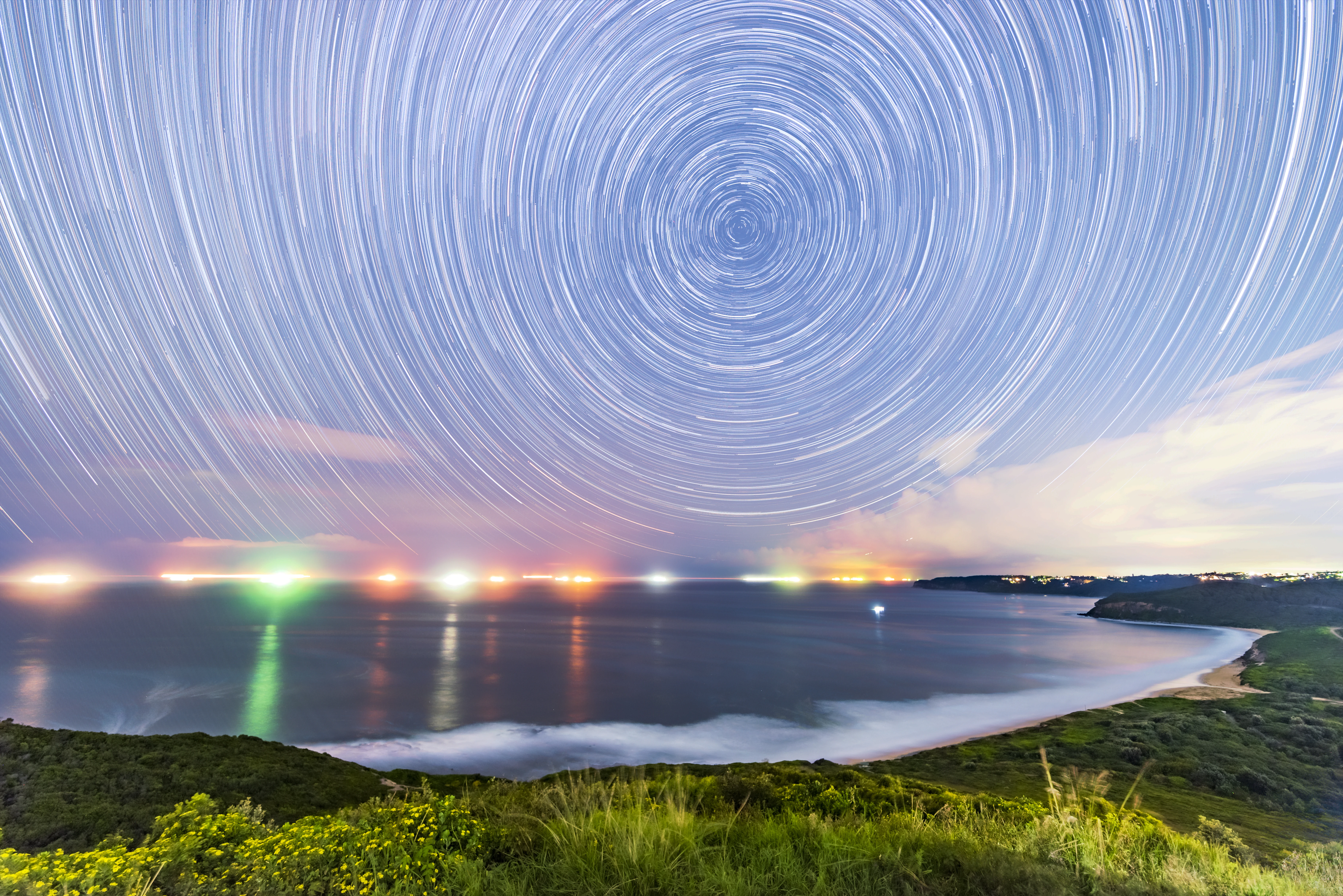 A night landscape view of a coastal beach with long exposure star trails swirling in the sky above the horizon; distant ship lights reflect on the water.