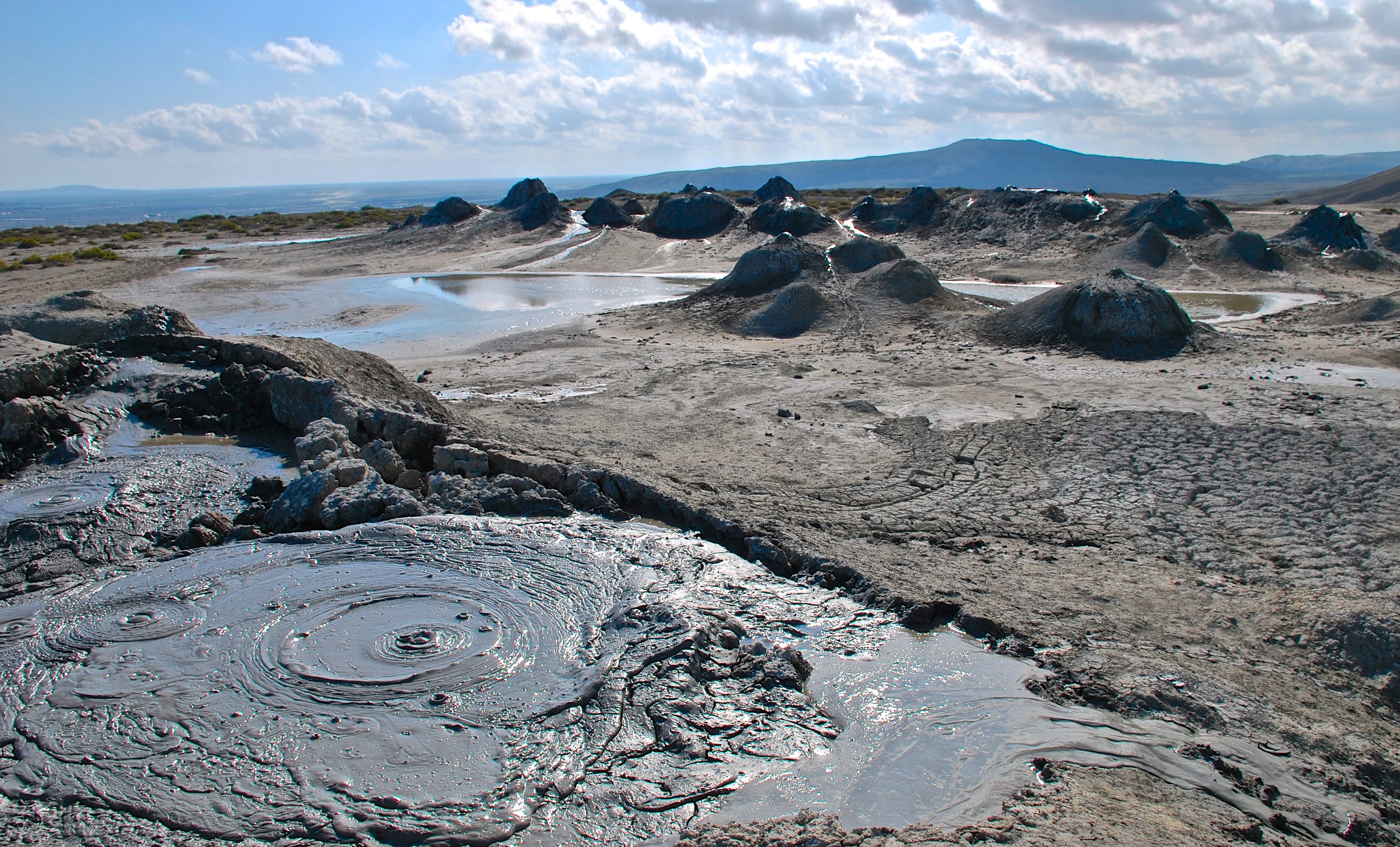 Una toma gran angular de un paisaje de barro gris y agrietado con múltiples volcanes de lodo burbujeantes y pequeños montículos bajo un cielo azul brillante.