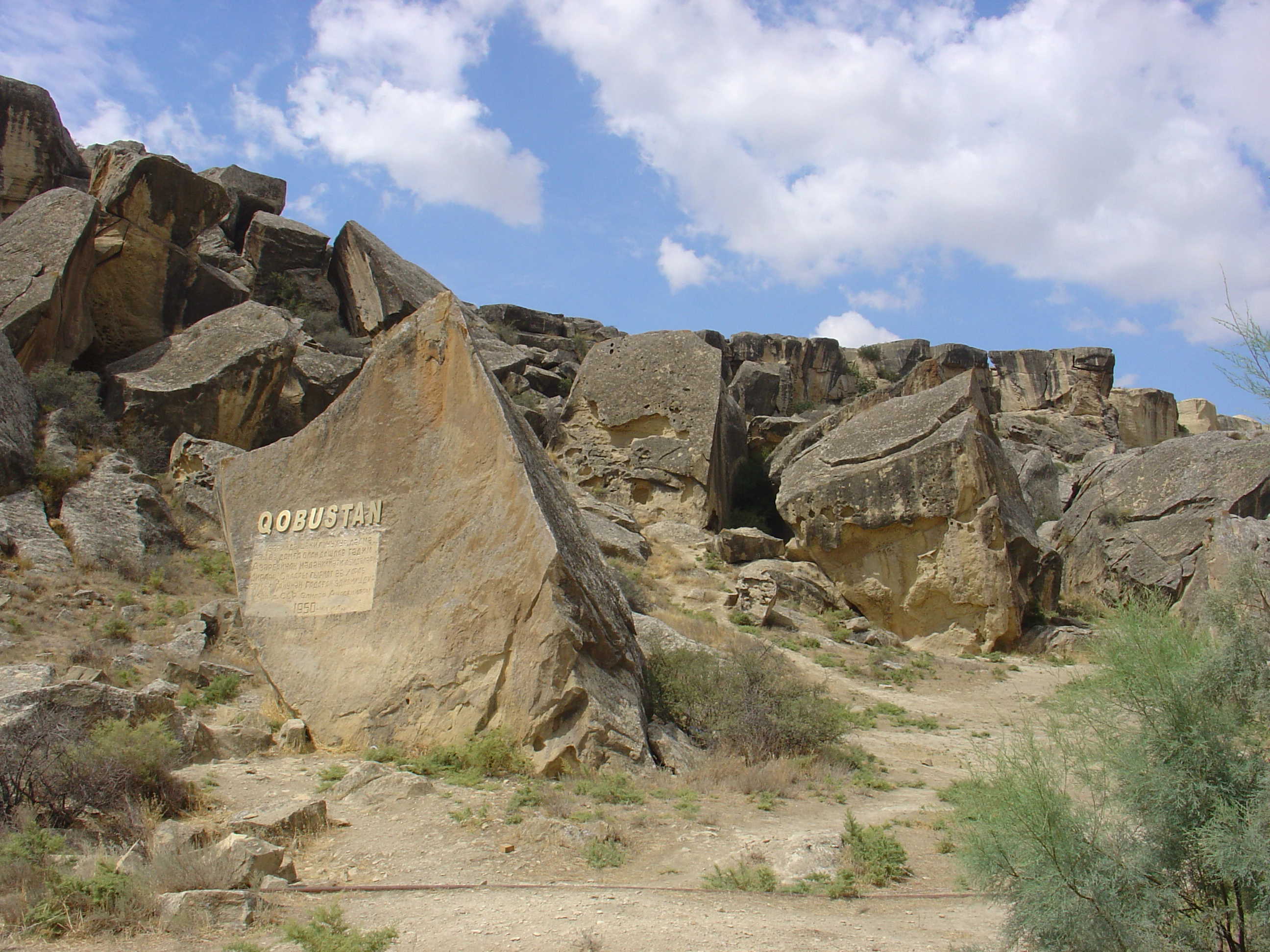Una gran roca triangular con la palabra QOBUSTAN grabada, situada en la entrada de un paisaje rocoso y árido bajo un cielo azul.