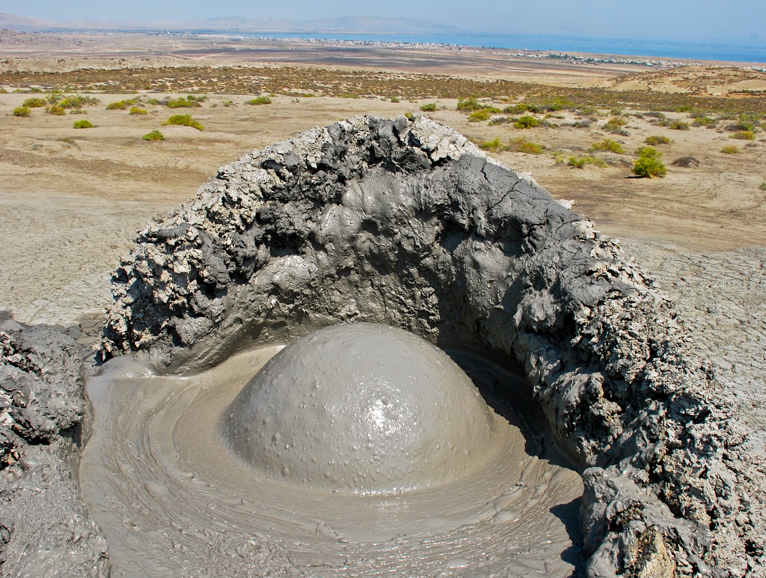 Un gran burbujeo de un volcán de lodo gris que erupciona en un paisaje desértico con vegetación escasa bajo un cielo brillante.
