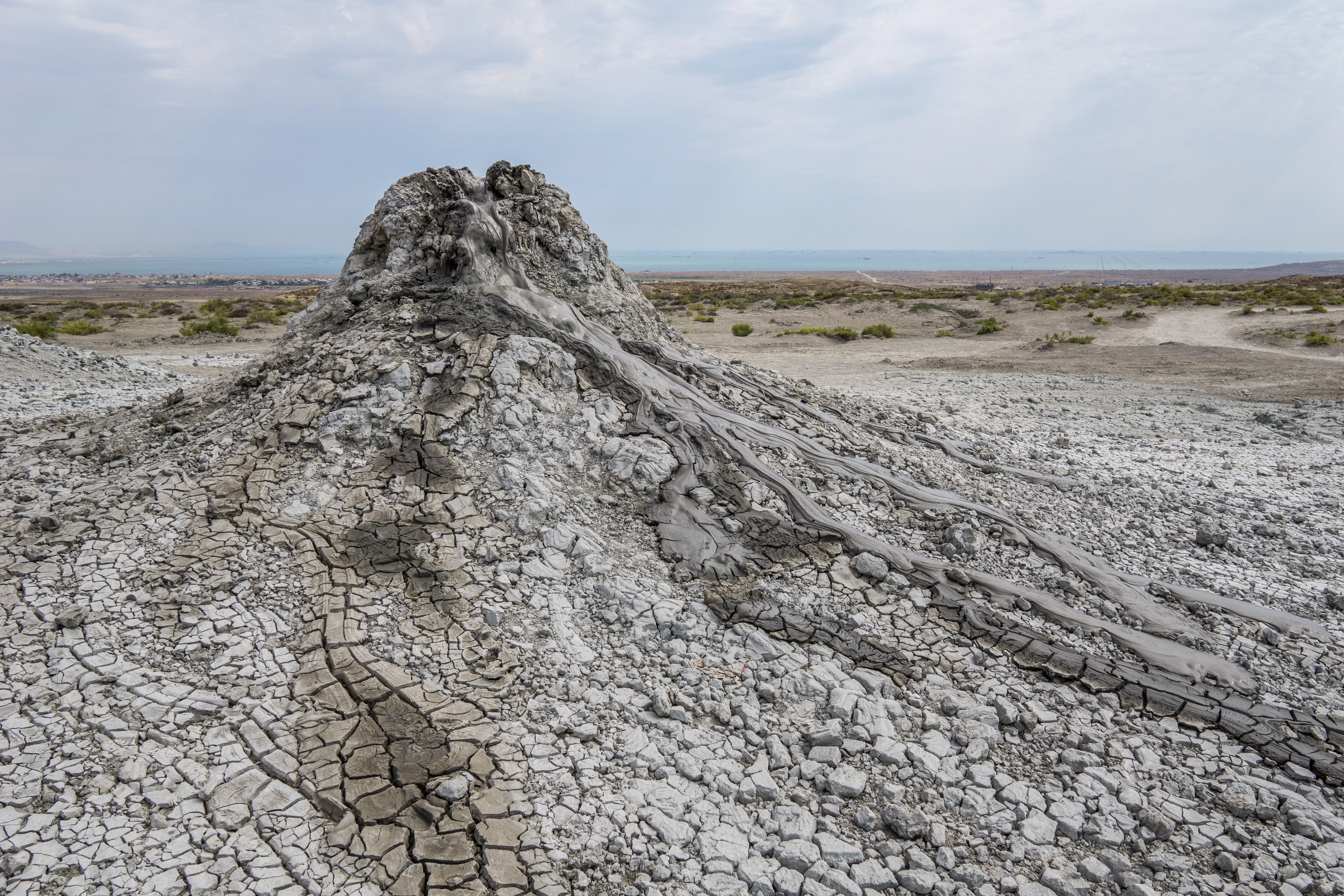 Un pequeño volcán de lodo cónico, con barro gris, agrietado y seco cubriendo sus laderas y el terreno llano y estéril circundante, bajo un cielo nublado.