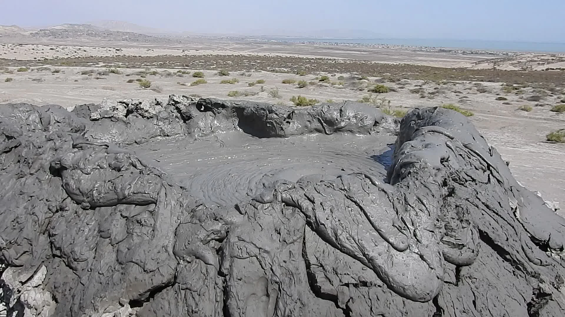 Una vista en primer plano de un cráter de volcán de lodo gris con barro burbujeante, enmarcado por un paisaje vasto, seco y árido con vegetación escasa.