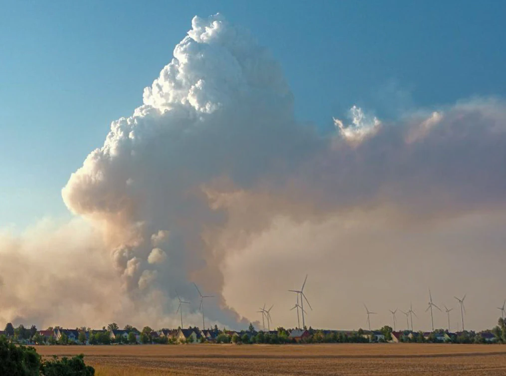 A massive plume of white and gray smoke rises into a clear blue sky above a flat horizon with wind turbines and a small residential settlement in the distance.