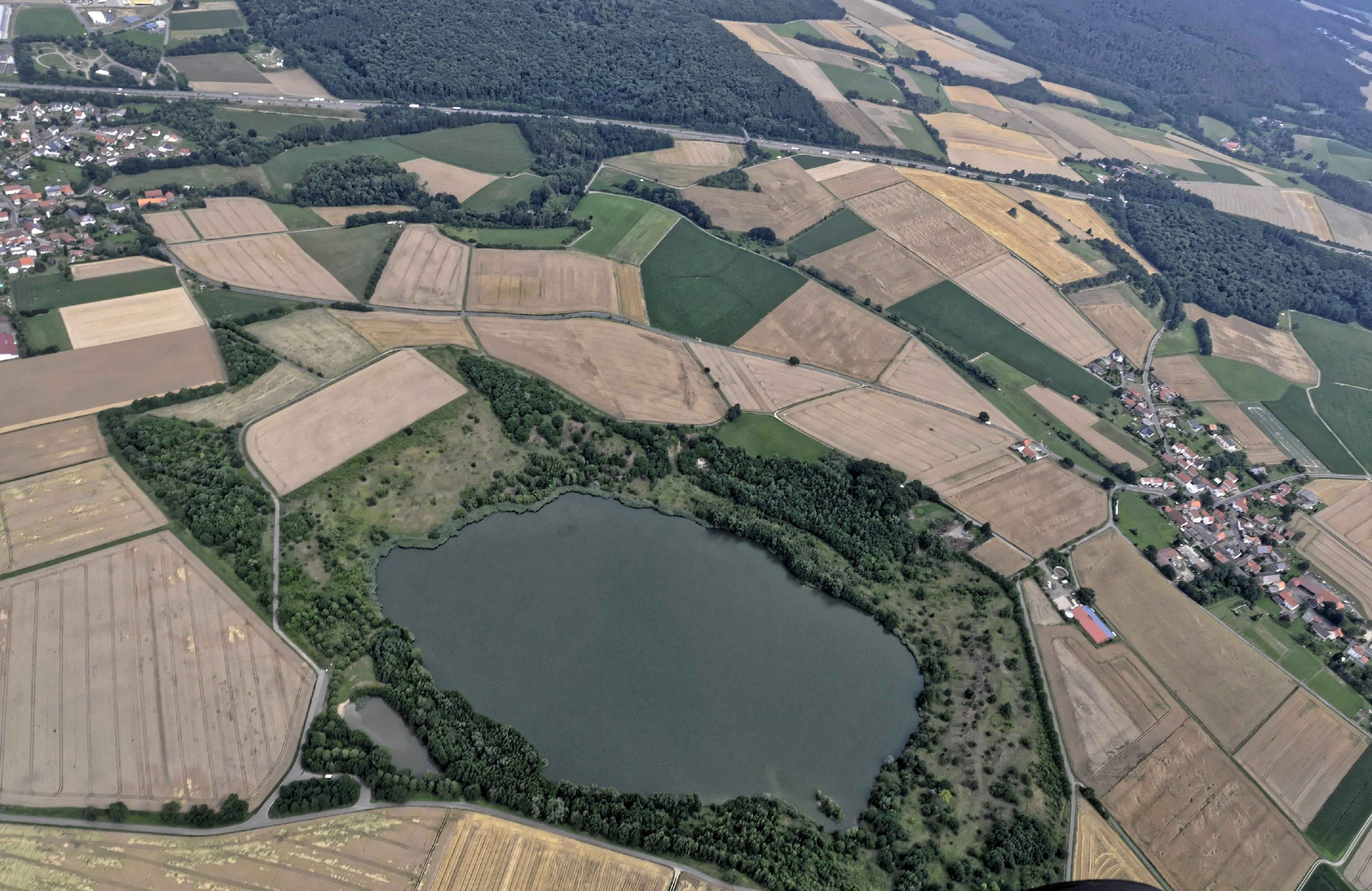 An aerial perspective of Goldbergsee, a circular lake surrounded by patches of forest and extensive agricultural fields, with residential areas visible at the edges.