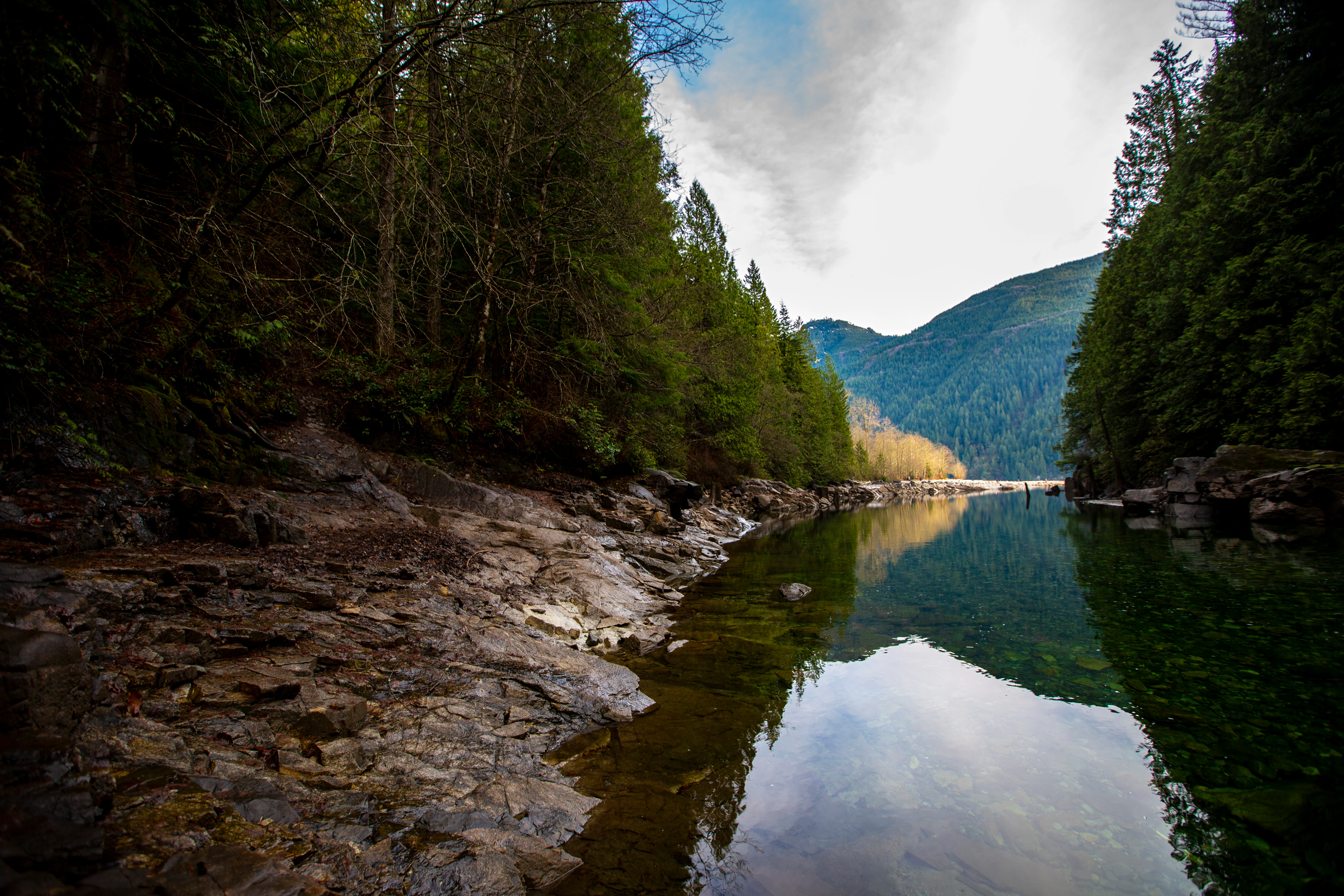 A wide angle view of a calm, reflective river flowing between steep, rocky banks covered in dense evergreen trees.