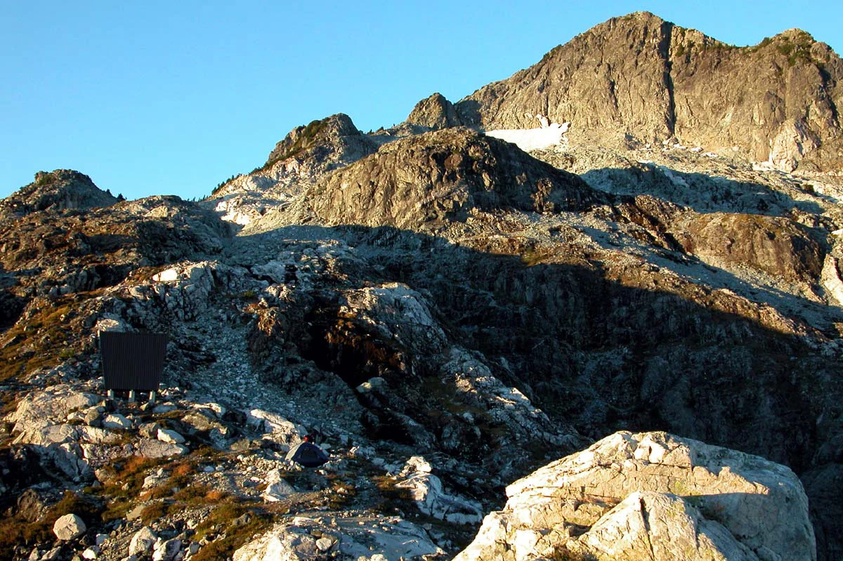Rugged, rocky alpine peak under clear blue sky with a small emergency shelter visible on the lower left ridge.