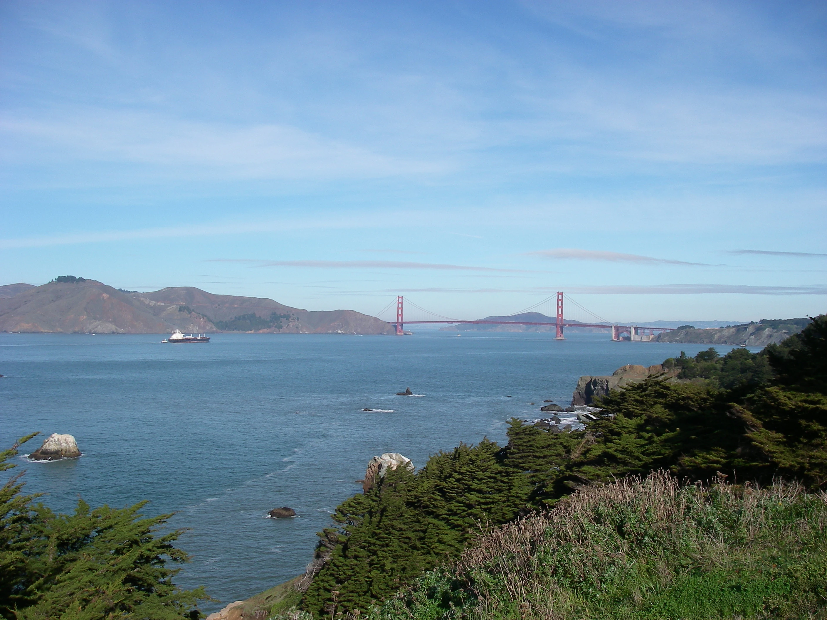 A wide coastal view showing the Golden Gate Bridge in the distance with the Lands End headlands in the foreground.
