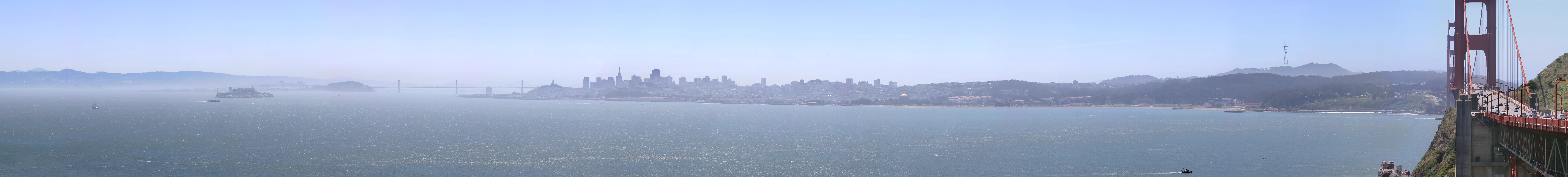 A wide panoramic photograph showing a vast expanse of the San Francisco Bay, with the partial structure of the Golden Gate Bridge on the right and the distant San Francisco skyline visible through a light haze.