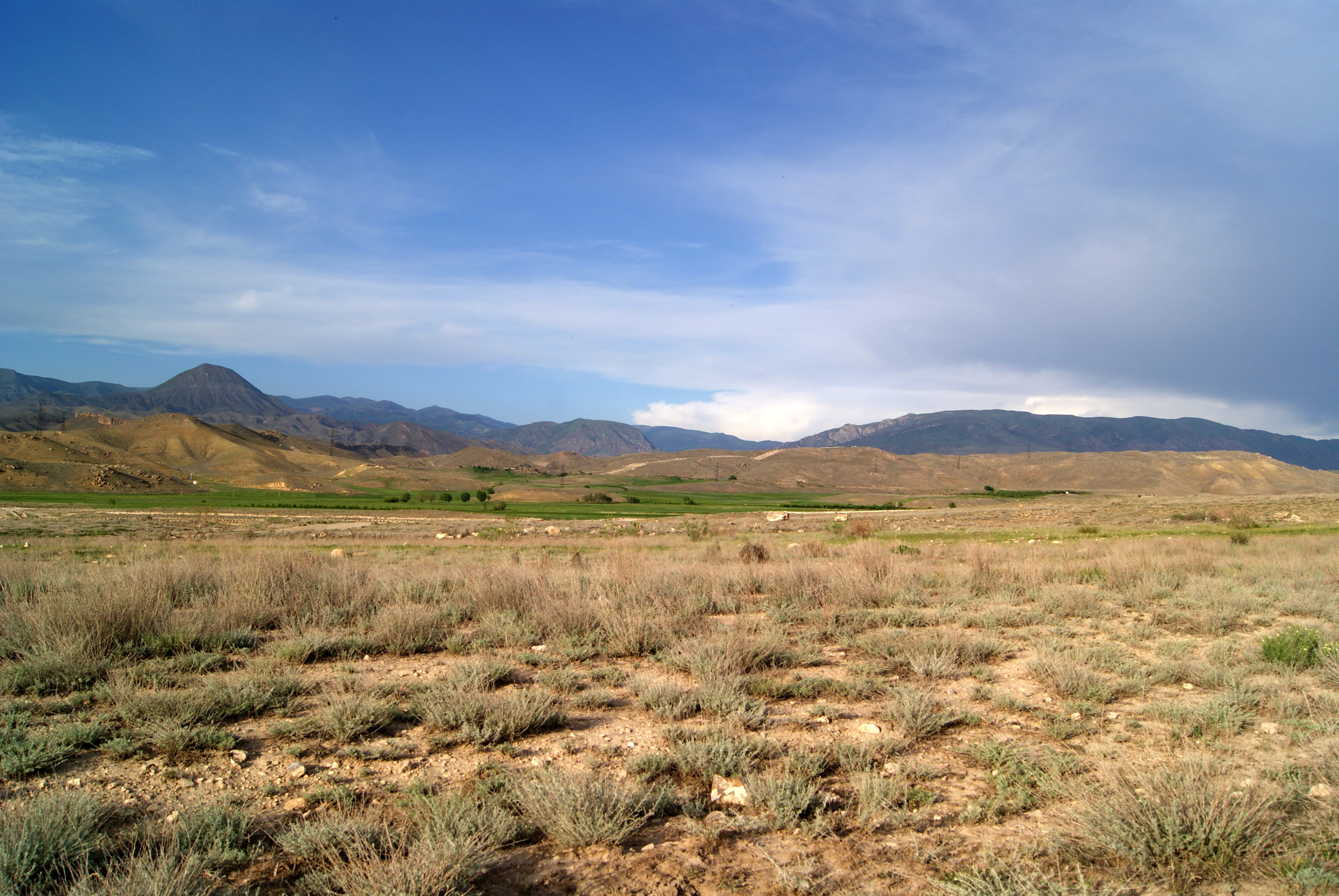 A wide, eye-level landscape shot of a dry, sandy semi-desert terrain with sparse shrubs and grasses in the foreground, leading to green fields and a mountain range under a blue sky with light clouds.