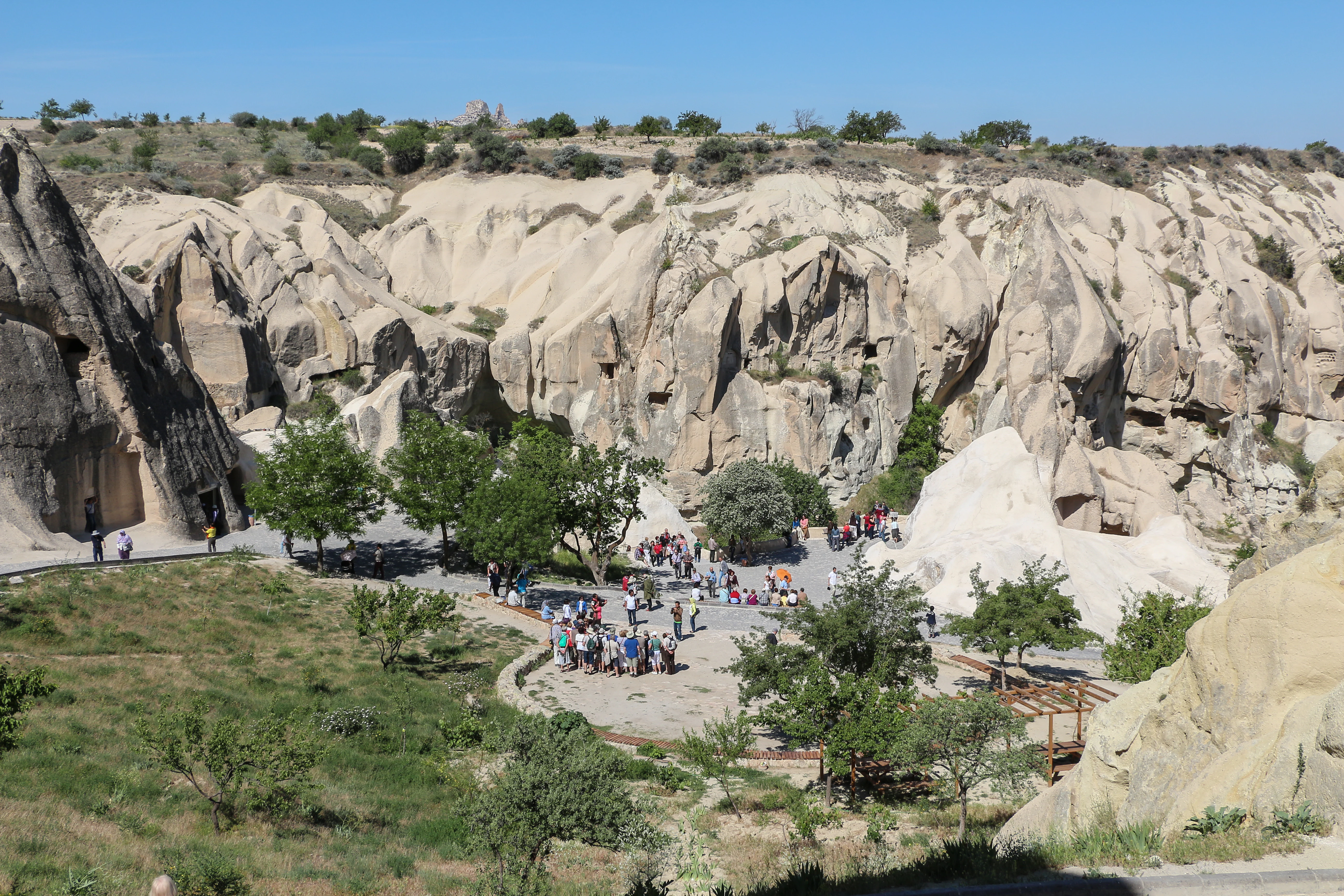 A wide view of Göreme Historical National Park featuring eroded rock formations, scattered trees, and a group of visitors on a pathway under a clear blue sky.