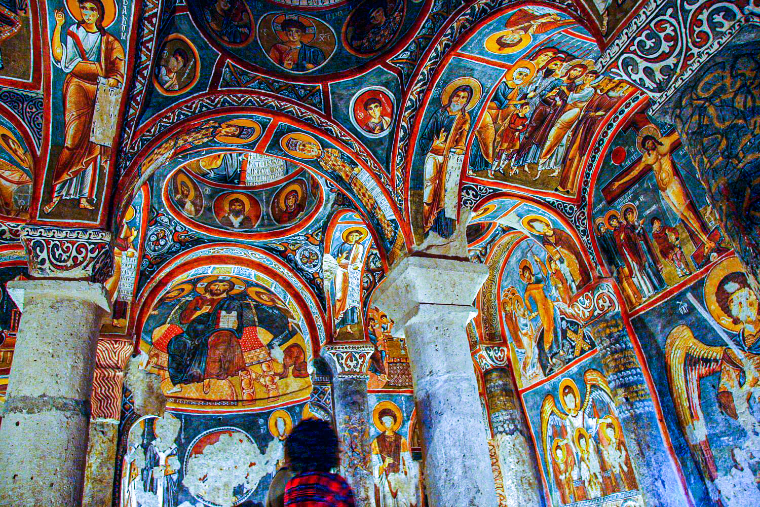 Interior of a historic church with colorful religious frescoes depicting figures and scenes on walls and ceilings, stone columns, and a visitor in the foreground