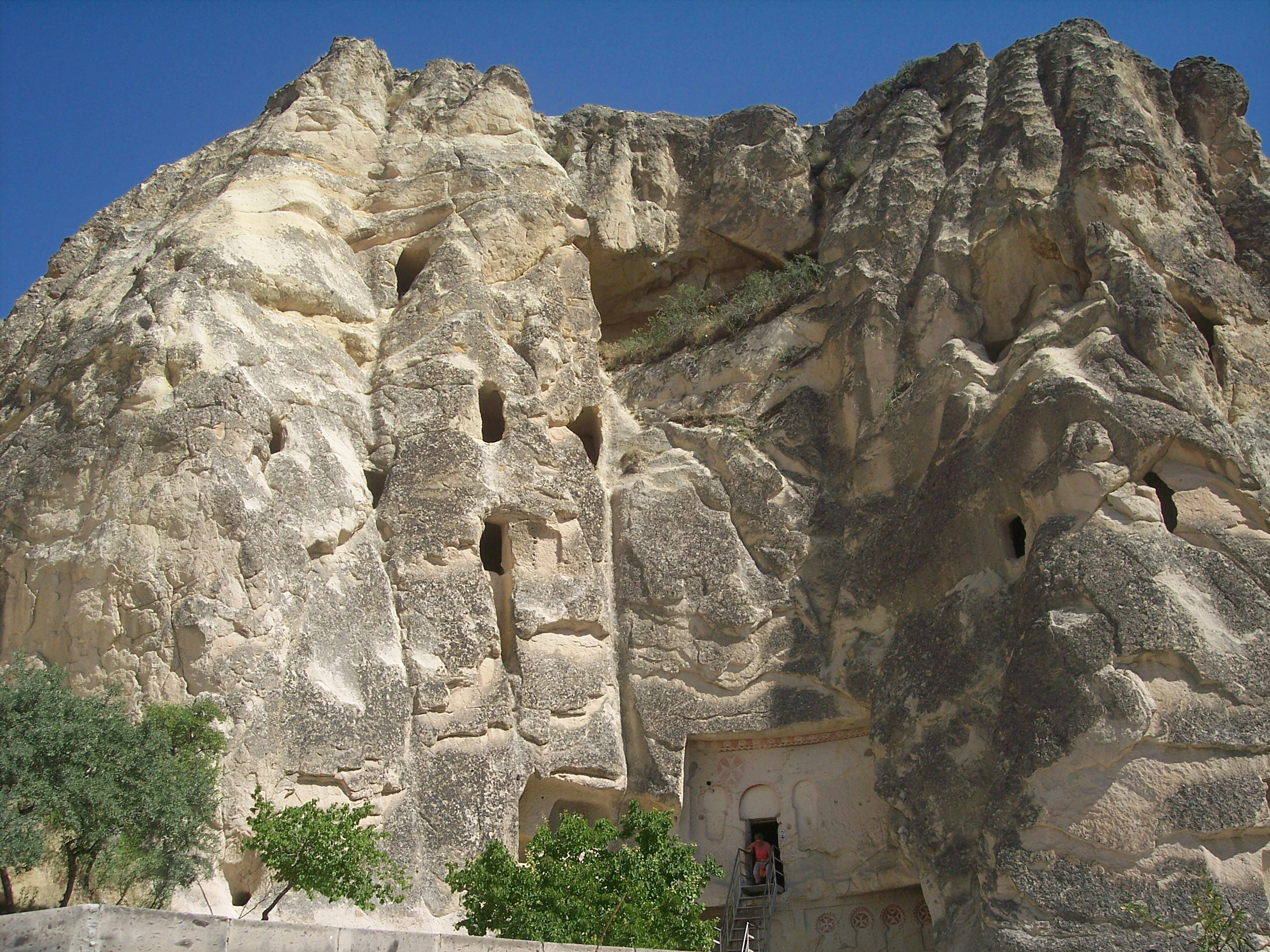 Rock face with multiple cave openings including an entrance to a church with a person standing at the doorway, trees in foreground, clear blue sky