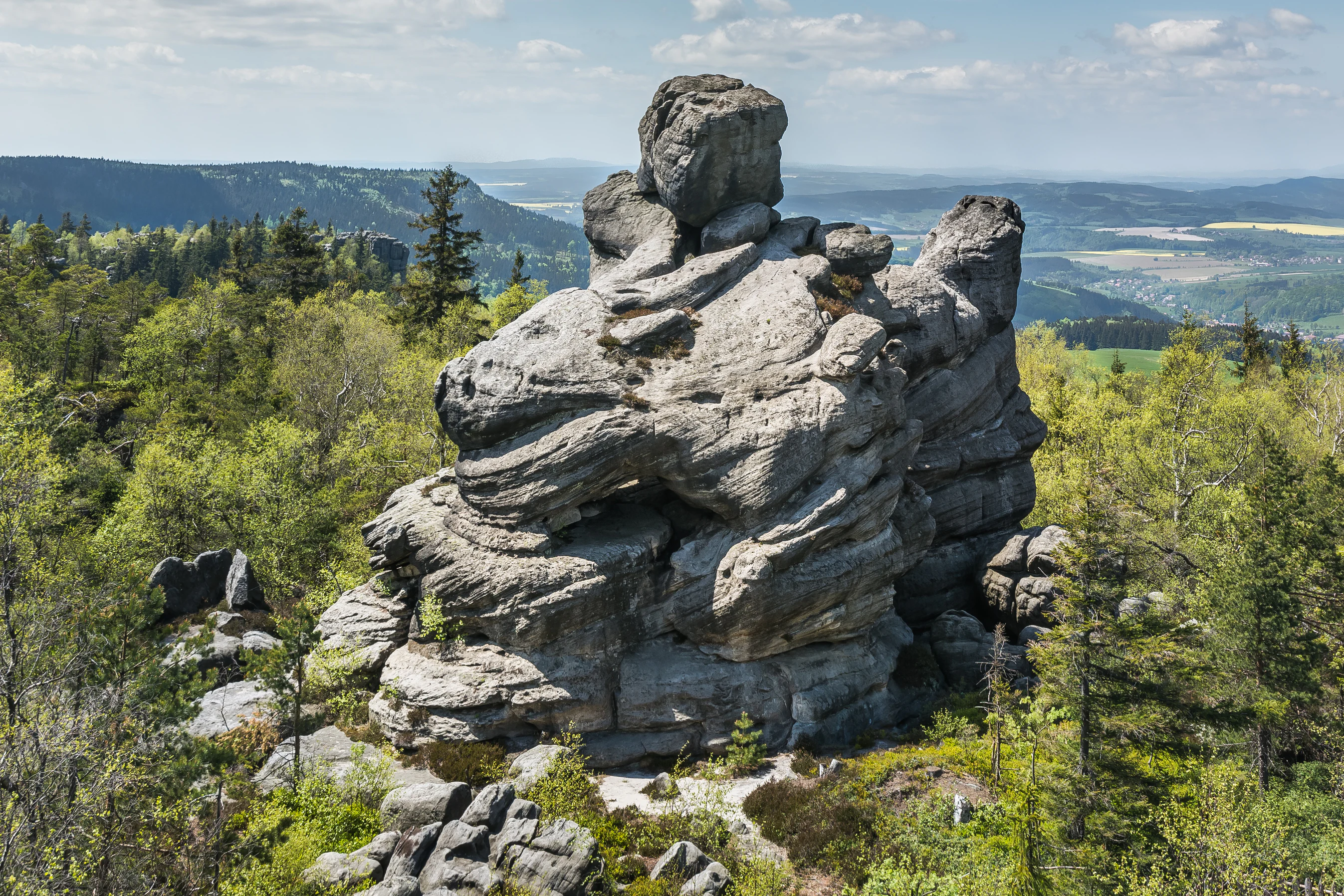 A large, weathered sandstone rock formation with distinct layered horizontal strata surrounded by a green forest and distant valley views.