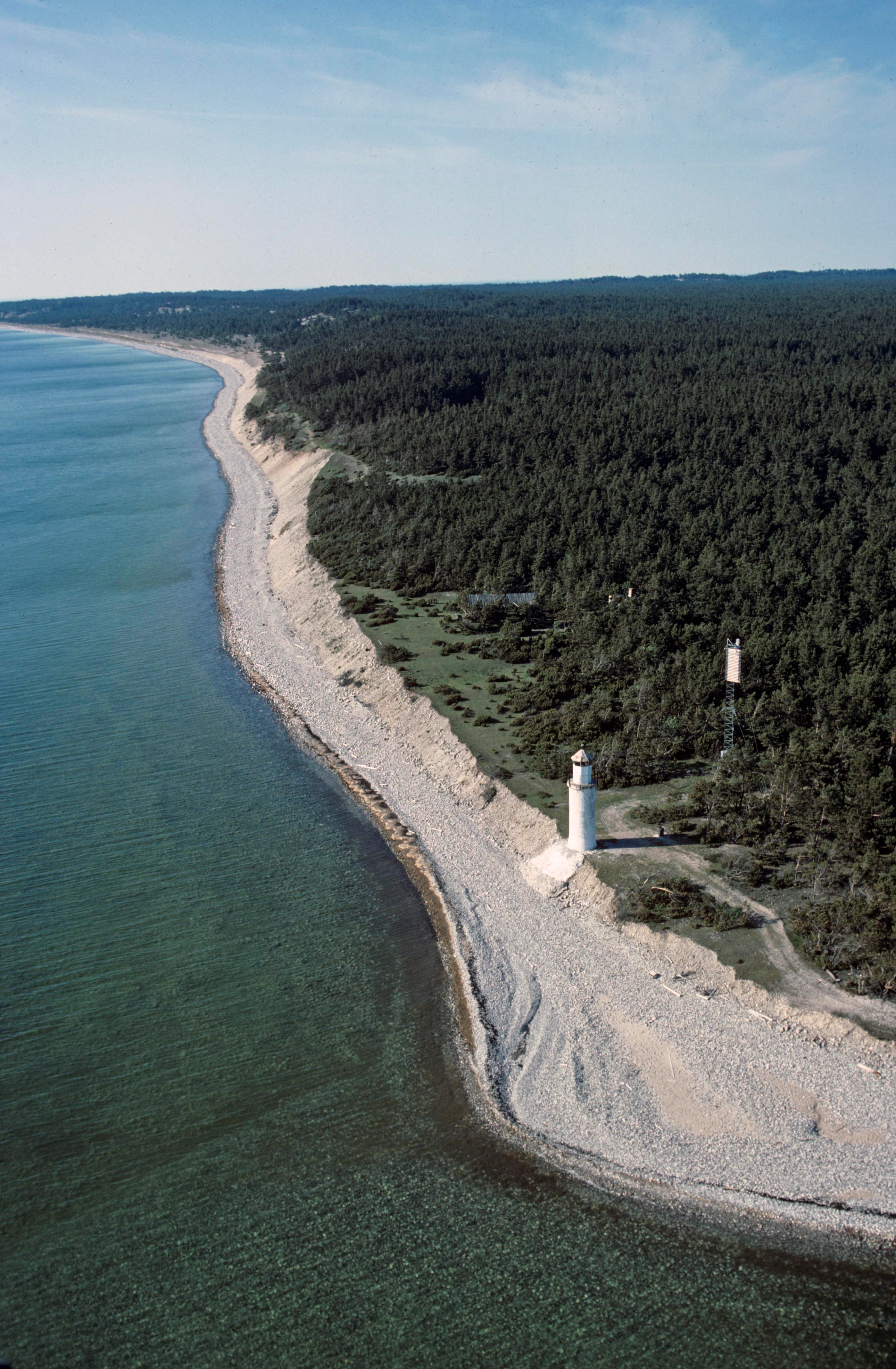 Aerial view of rocky coastline with white lighthouse, adjacent forested area, and calm sea under clear sky