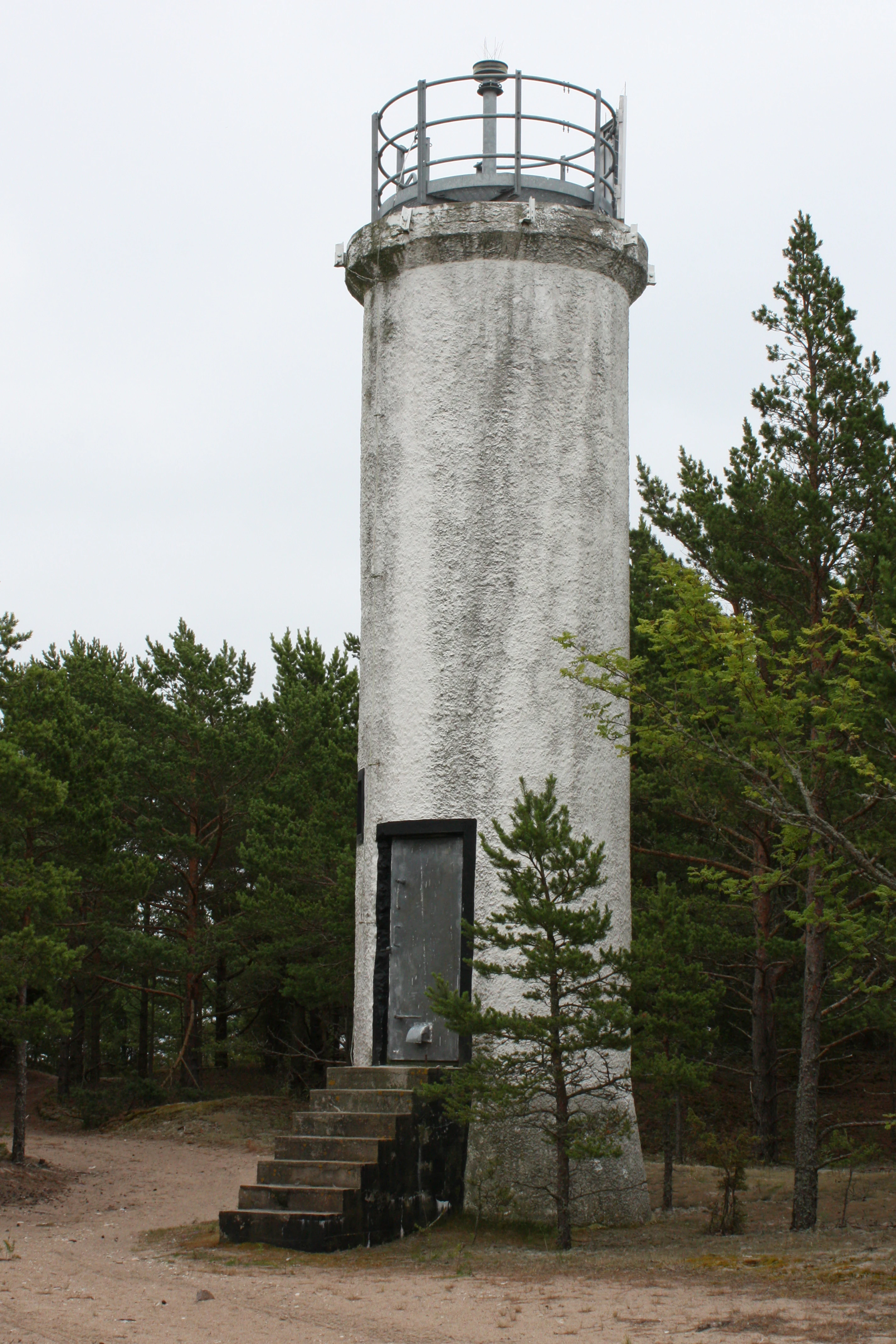 Weathered white lighthouse with metal railing, steps leading to entrance, surrounded by pine trees and sandy ground