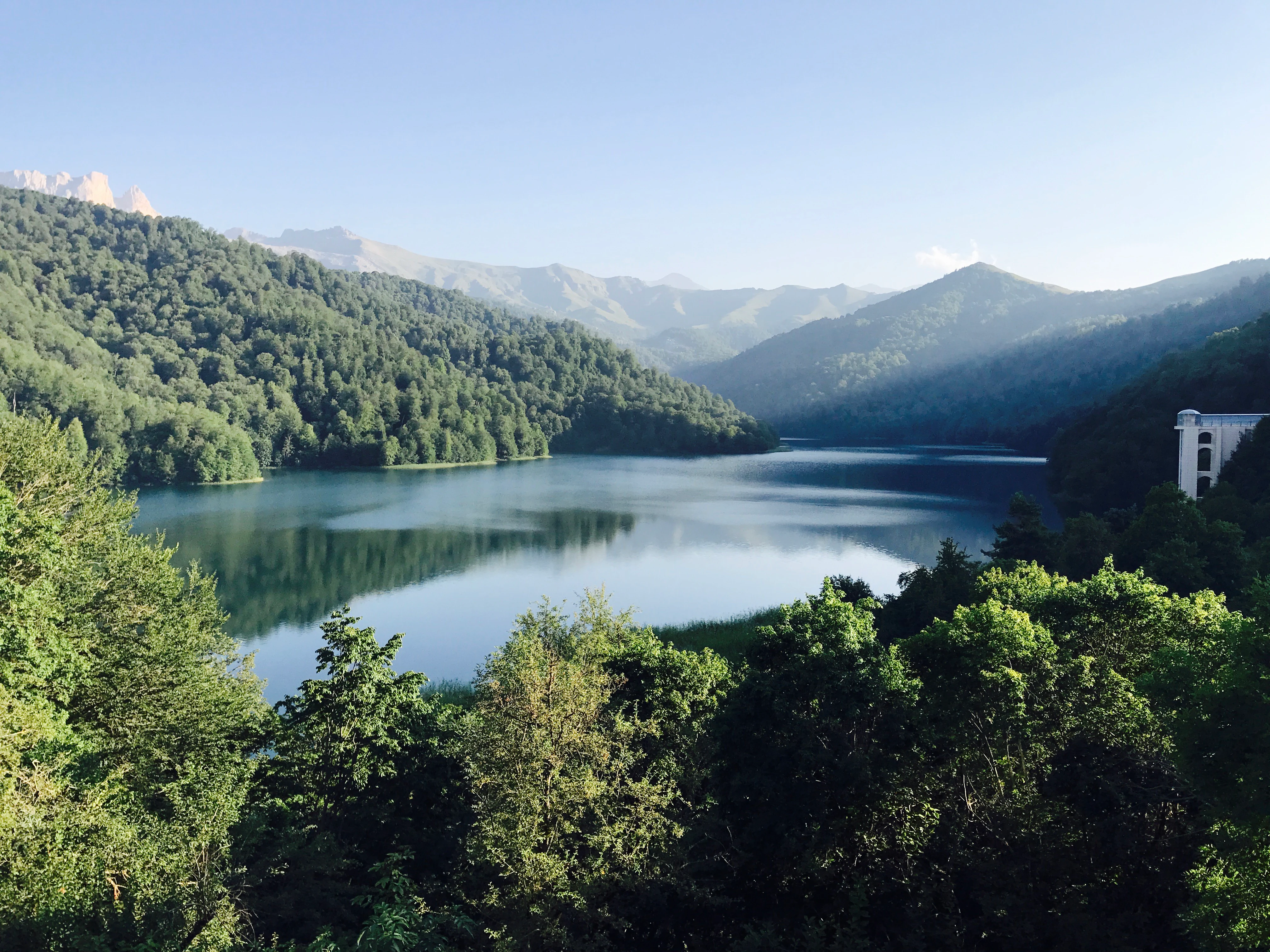 A serene lake reflecting surrounding mountains and dense green forests under a clear blue sky, with a small building visible on the right