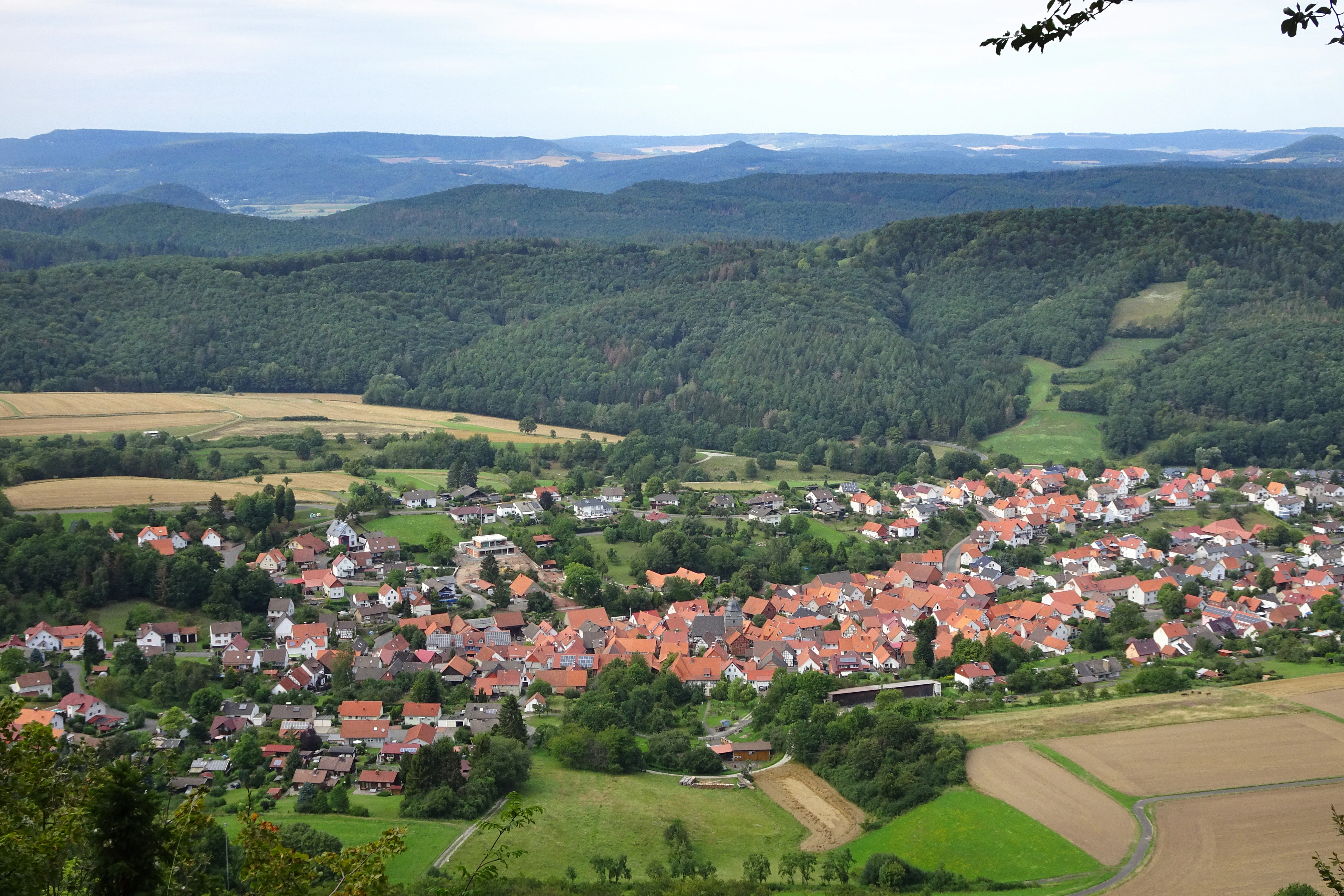 A high-angle landscape photograph showing a village with red-roofed houses in a valley, surrounded by green forests, agricultural fields, and rolling hills in the background.