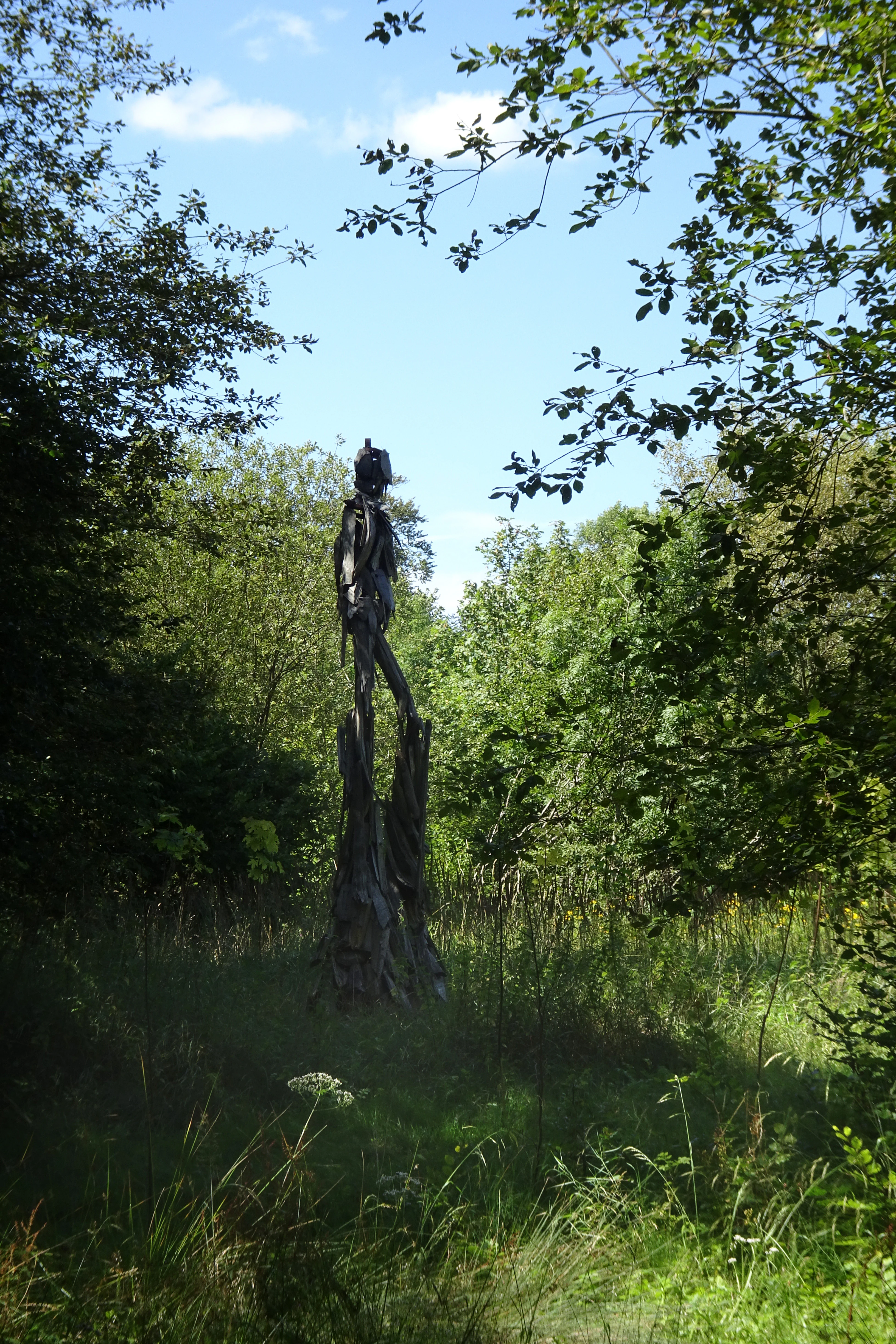 A tall, dark, slender sculpture of a human-like figure stands in a grassy clearing surrounded by green trees and a blue sky.