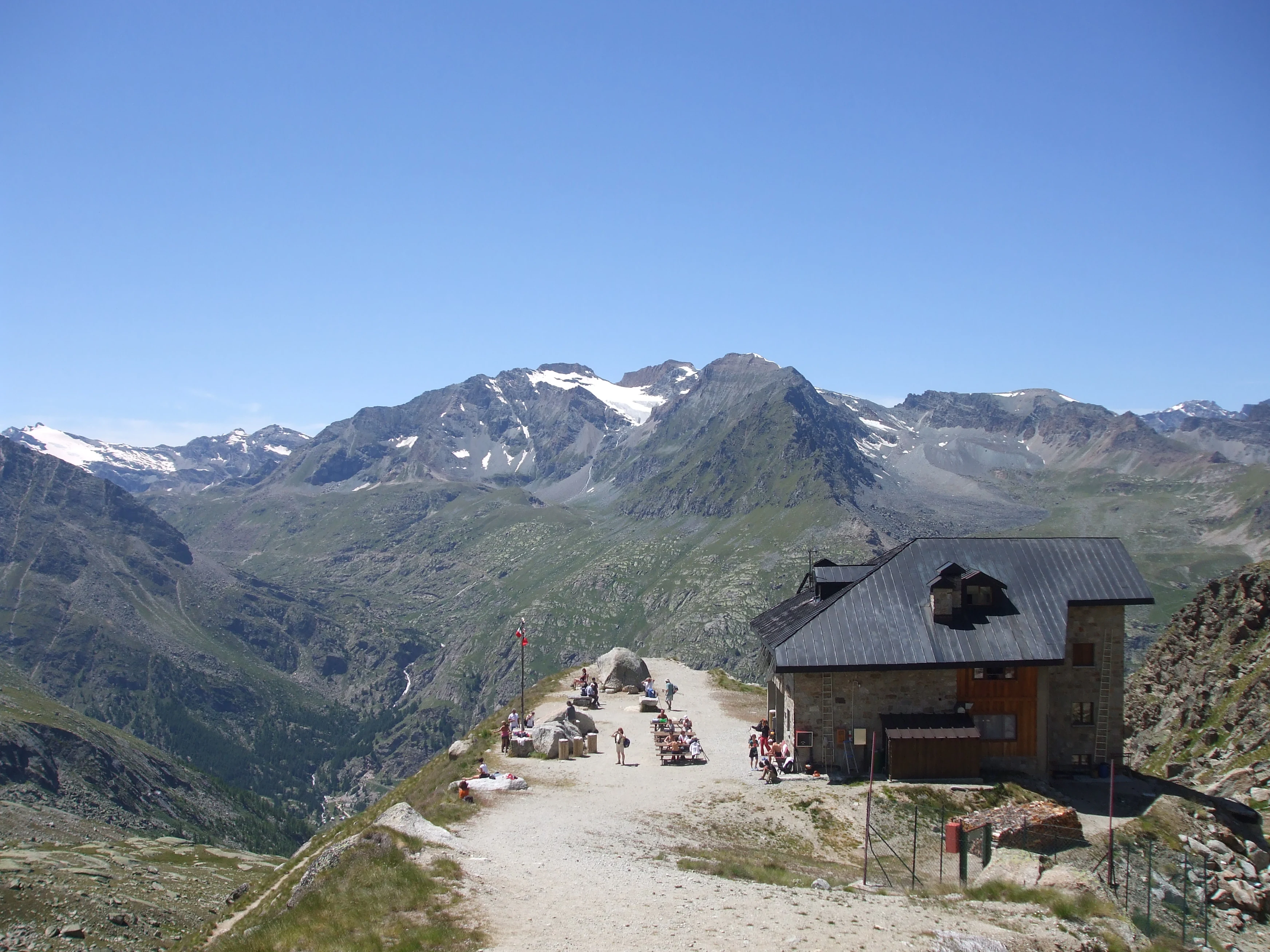 Mountain hut with people on a dirt path, mountain peaks with snow, and valley in background