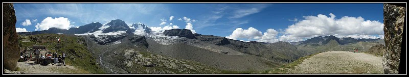 Panoramic landscape showing snow-capped mountain peaks, valleys, a dirt road with a vehicle, and stone structures on the left and right edges under a blue sky with scattered clouds
