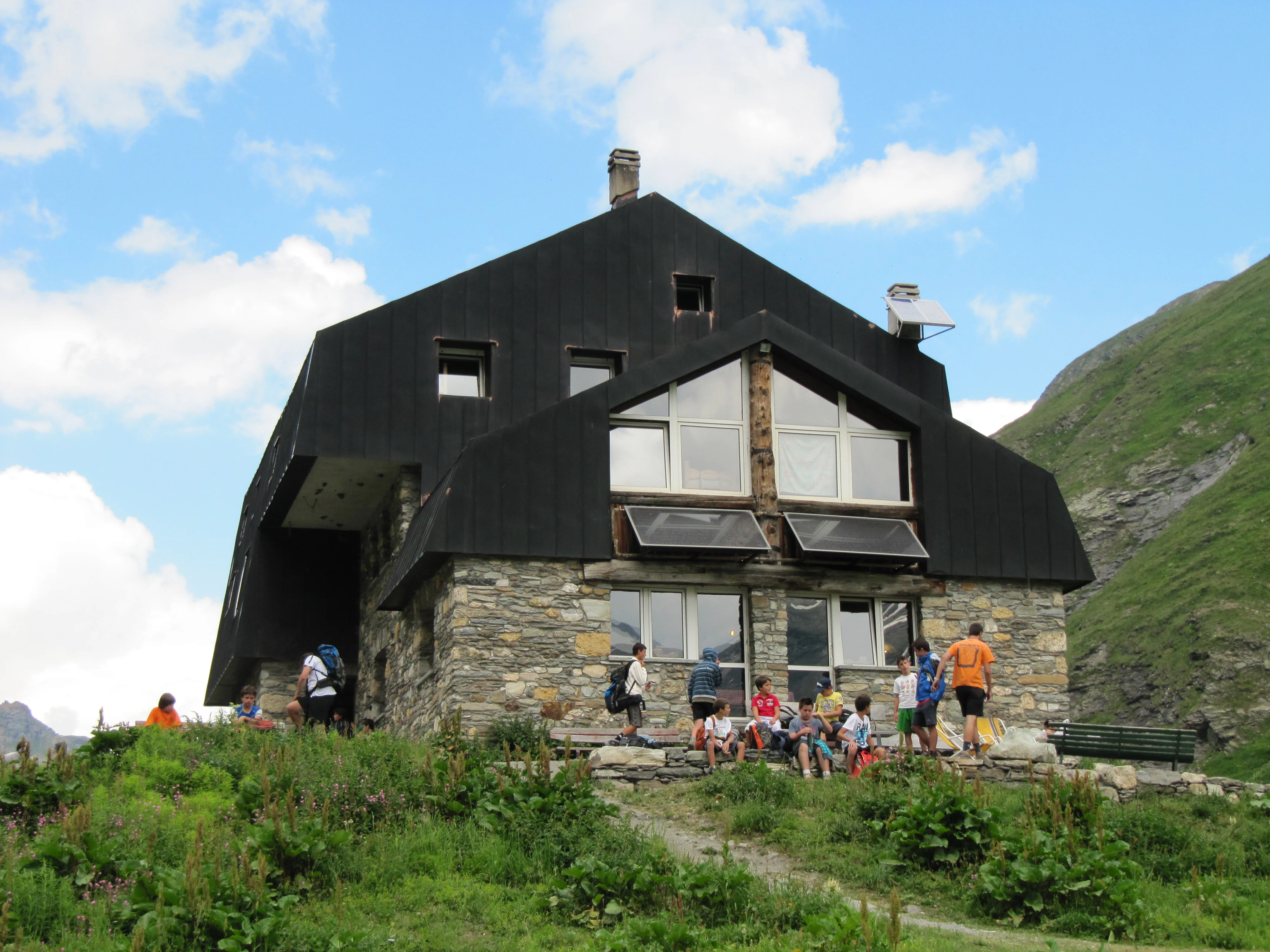 Stone mountain hut with black roof and several people gathered outside on a grassy area, with mountains in the background