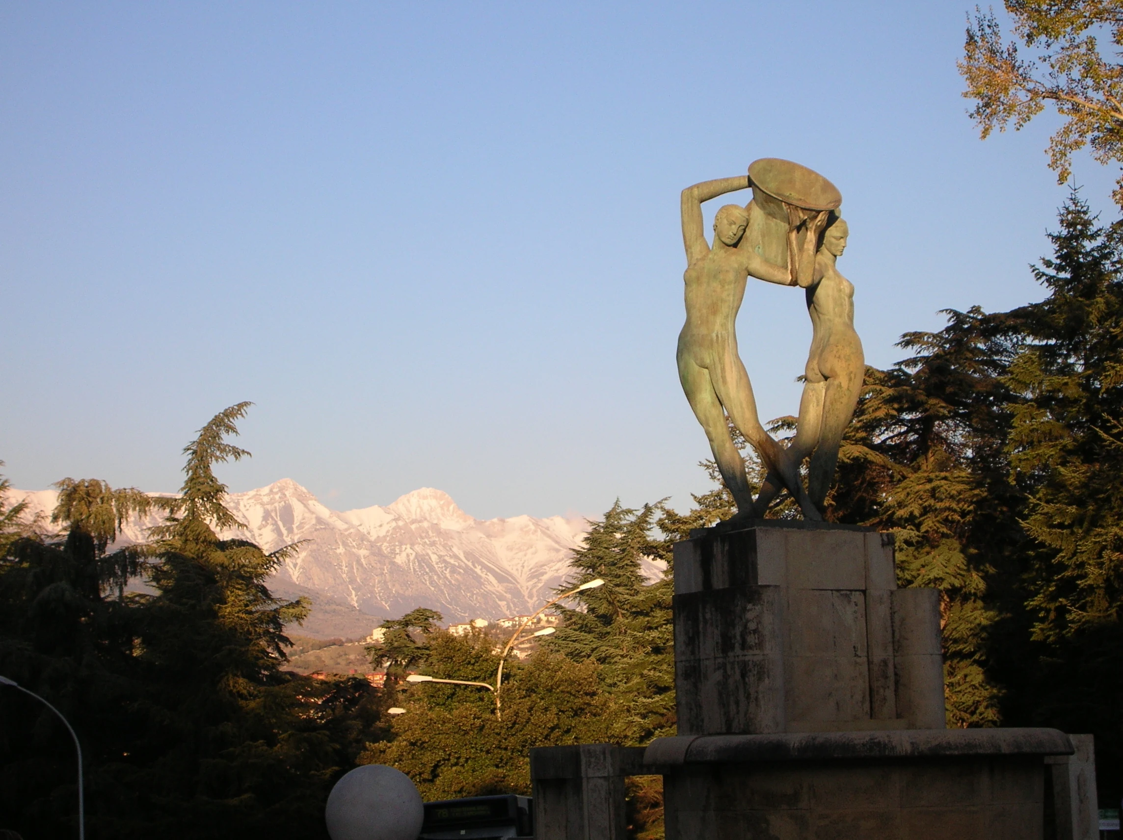 Bronze sculpture of three figures on stone pedestal with snow-capped mountains in background, trees and clear sky