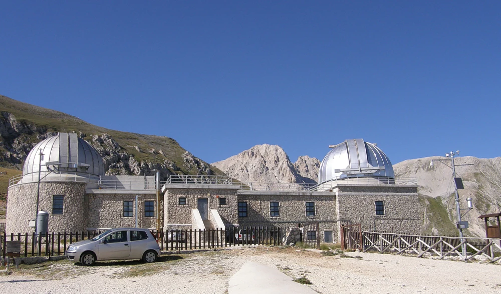 Stone building with two metallic domes, parked car, mountainous background under clear blue sky