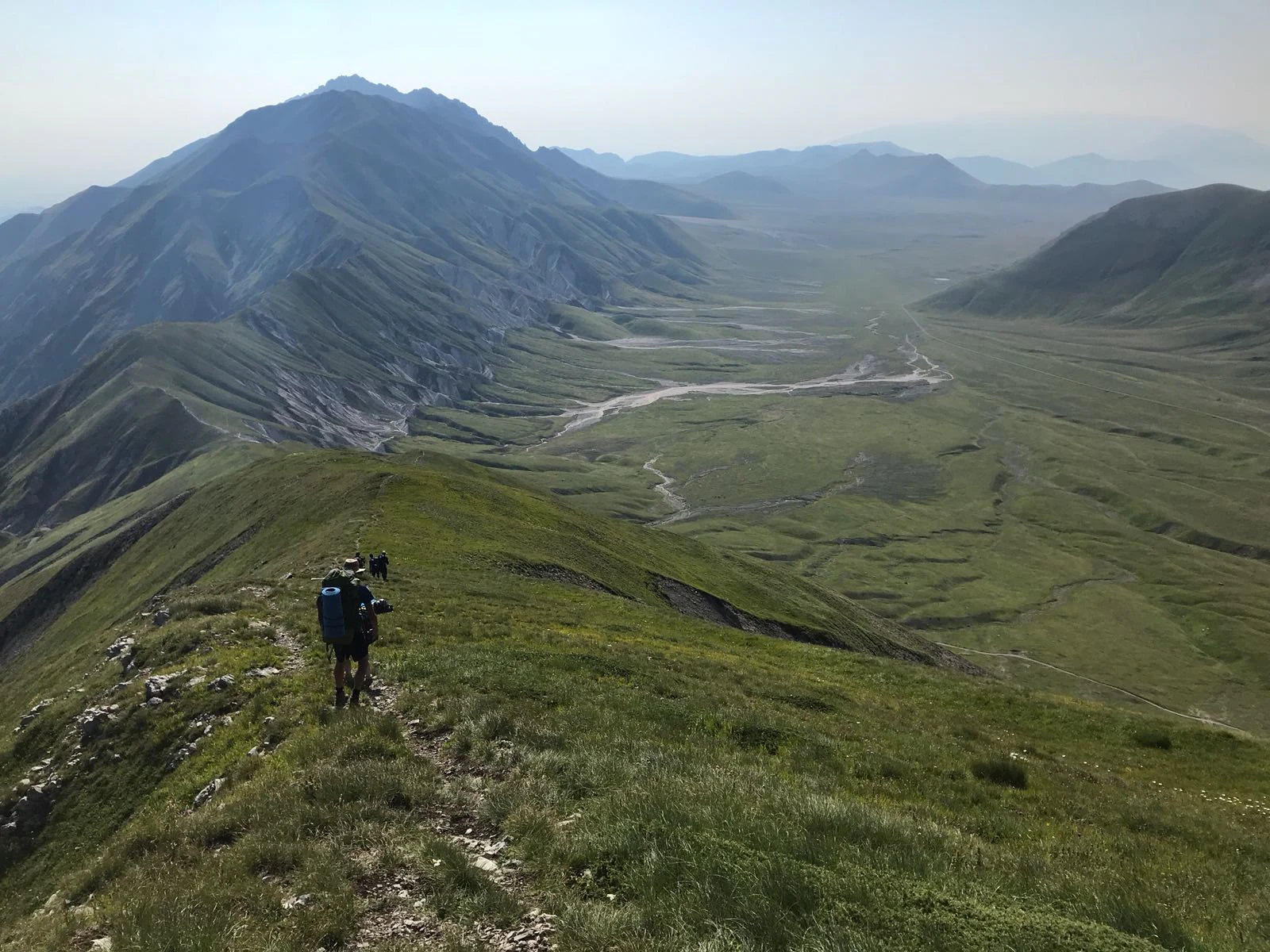 A hiker with a backpack walking on a rocky trail atop a grassy mountain ridge with a vast valley and distant mountain ranges under a clear sky