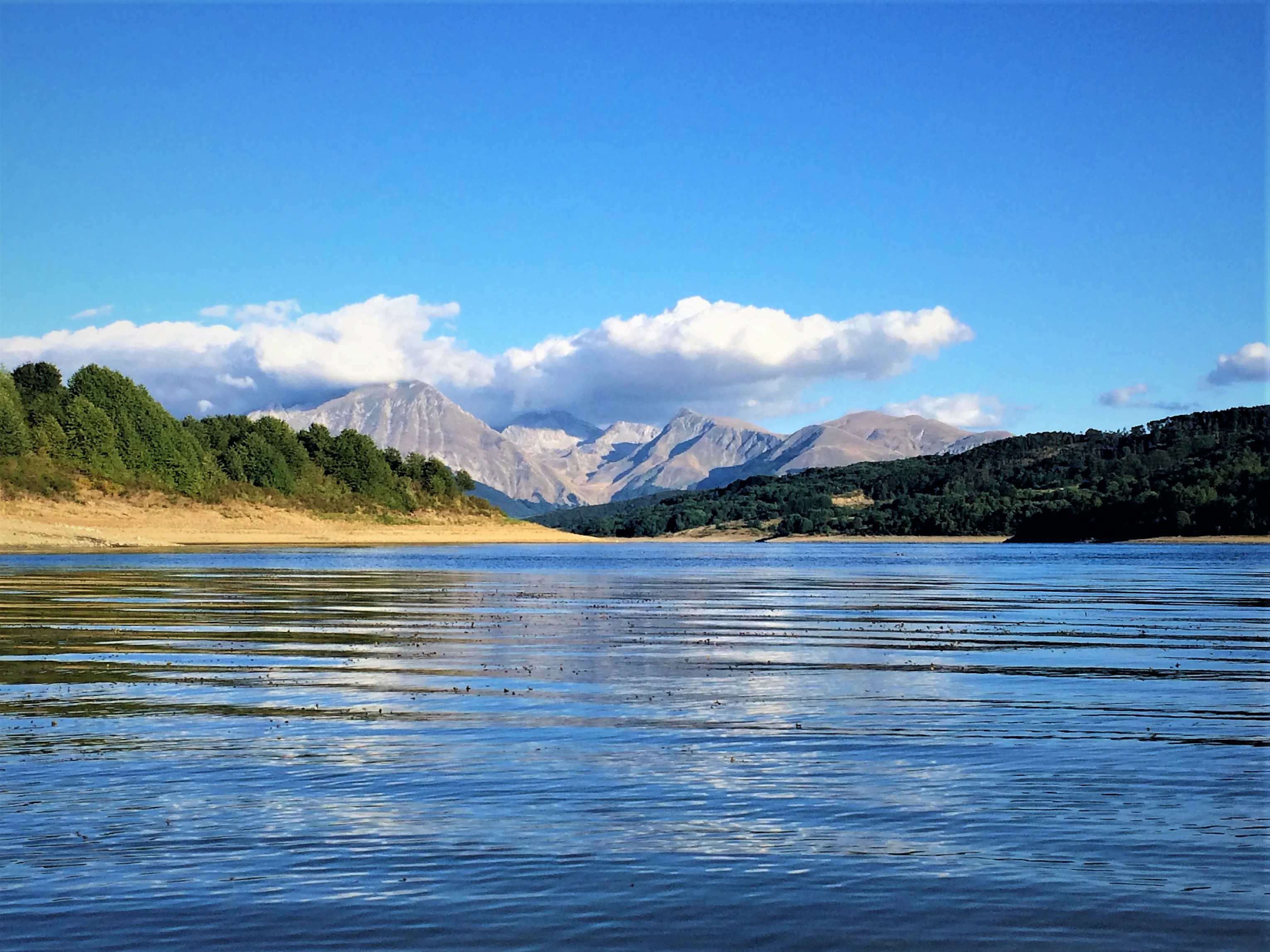 A calm lake reflecting blue sky and distant mountains, surrounded by forested hills