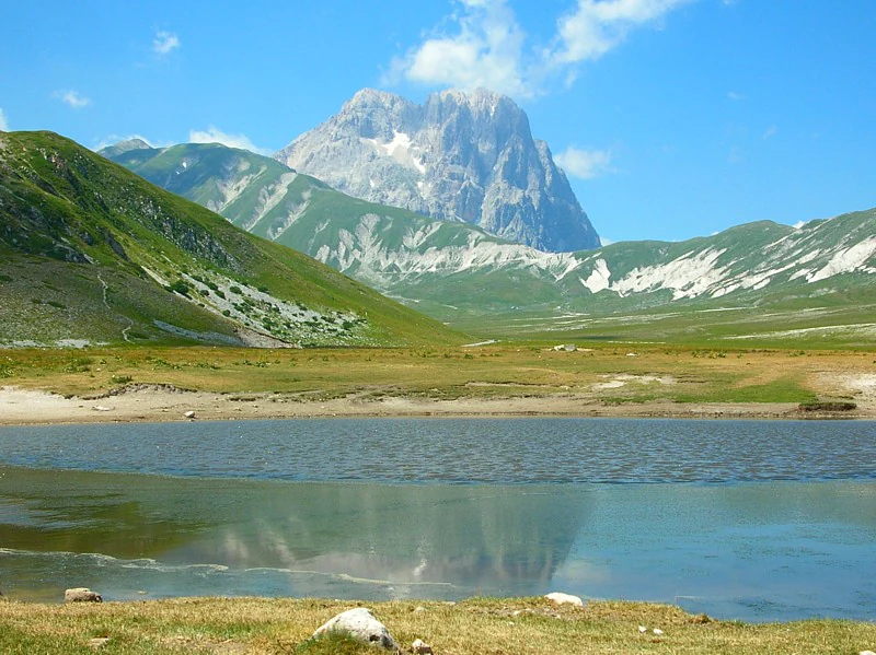 A lake reflecting a large mountain with green slopes under a clear blue sky