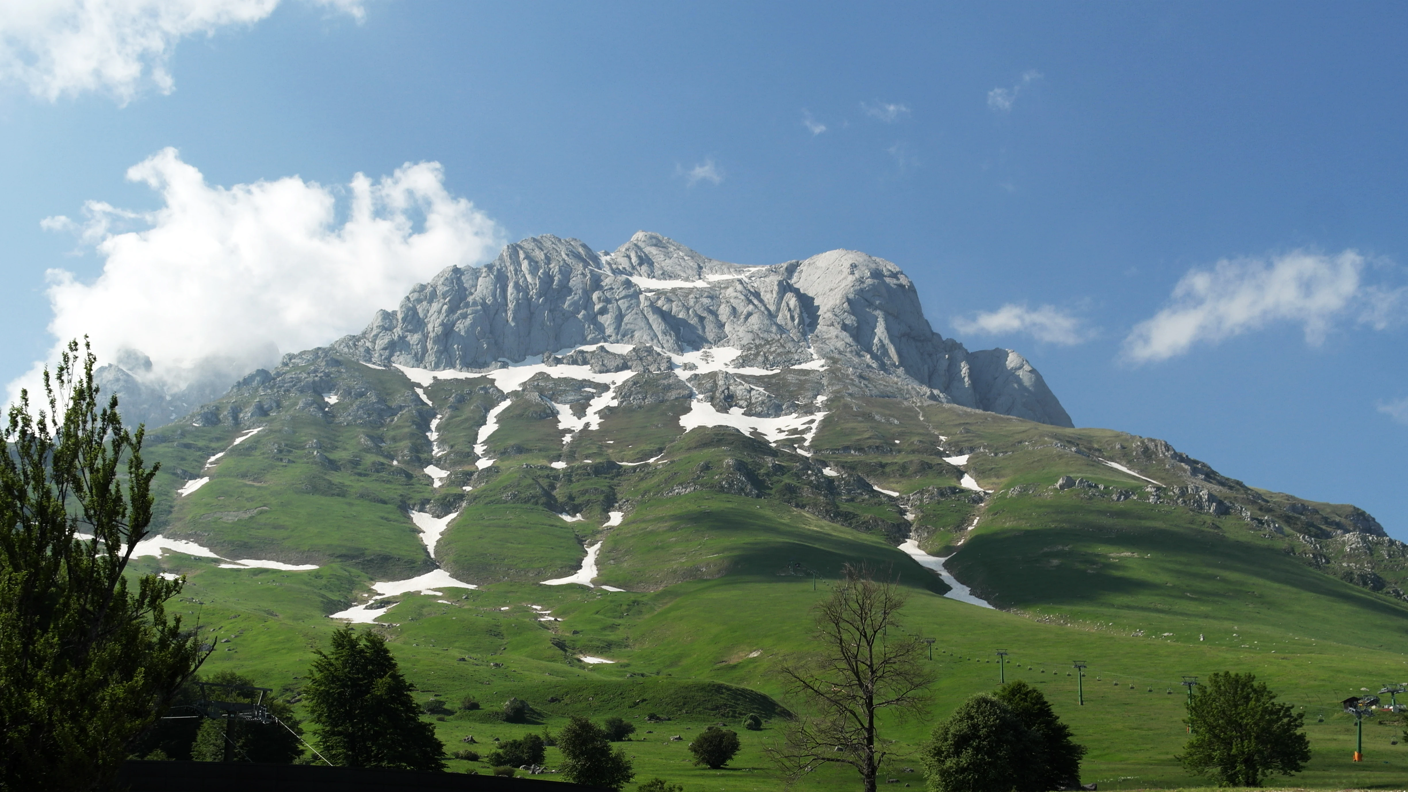 Mountain peak with snow patches on a green hillside, trees in the foreground and blue sky with clouds