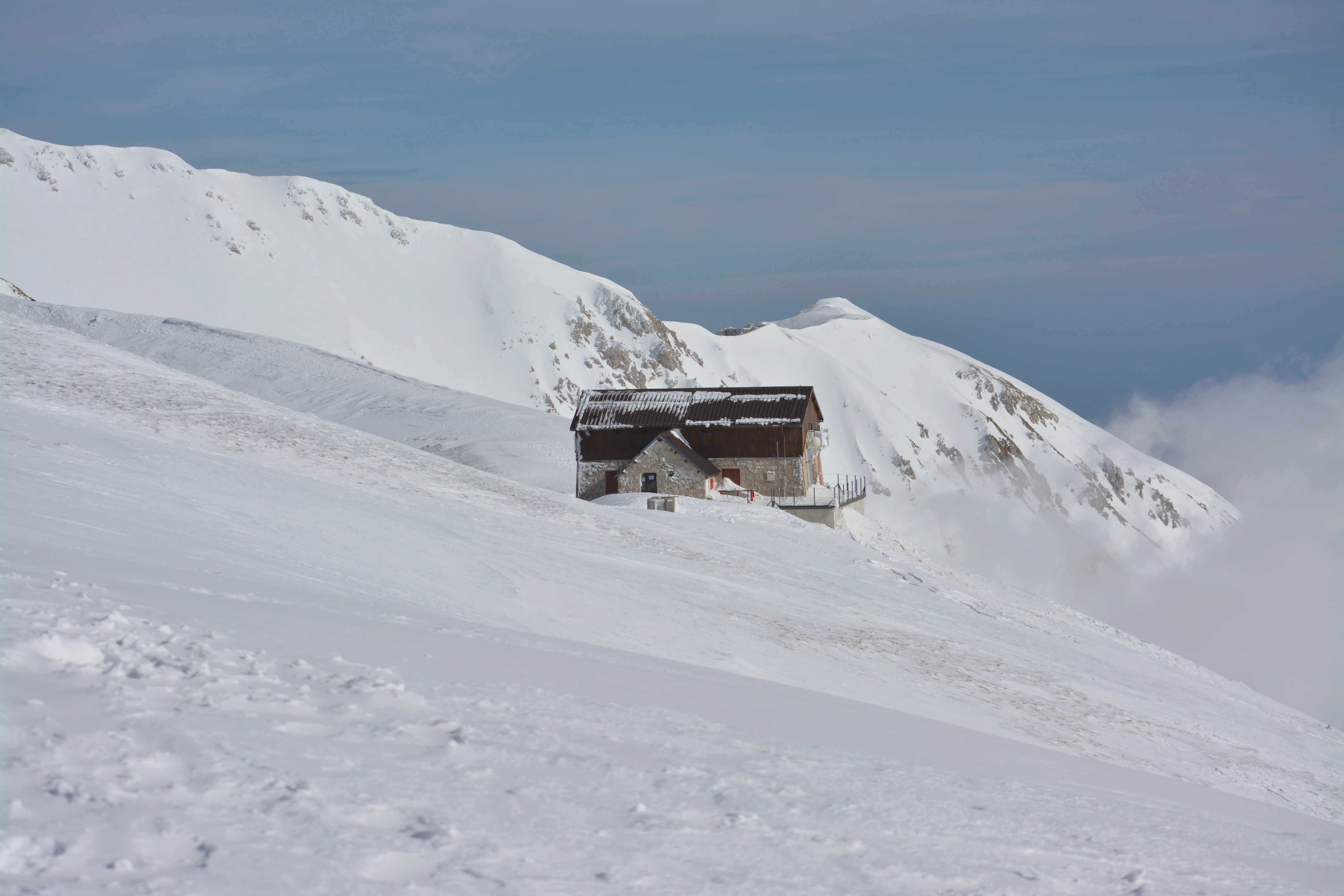 A wooden mountain hut situated on a snowy slope with snow-covered mountains in the background under a clear sky