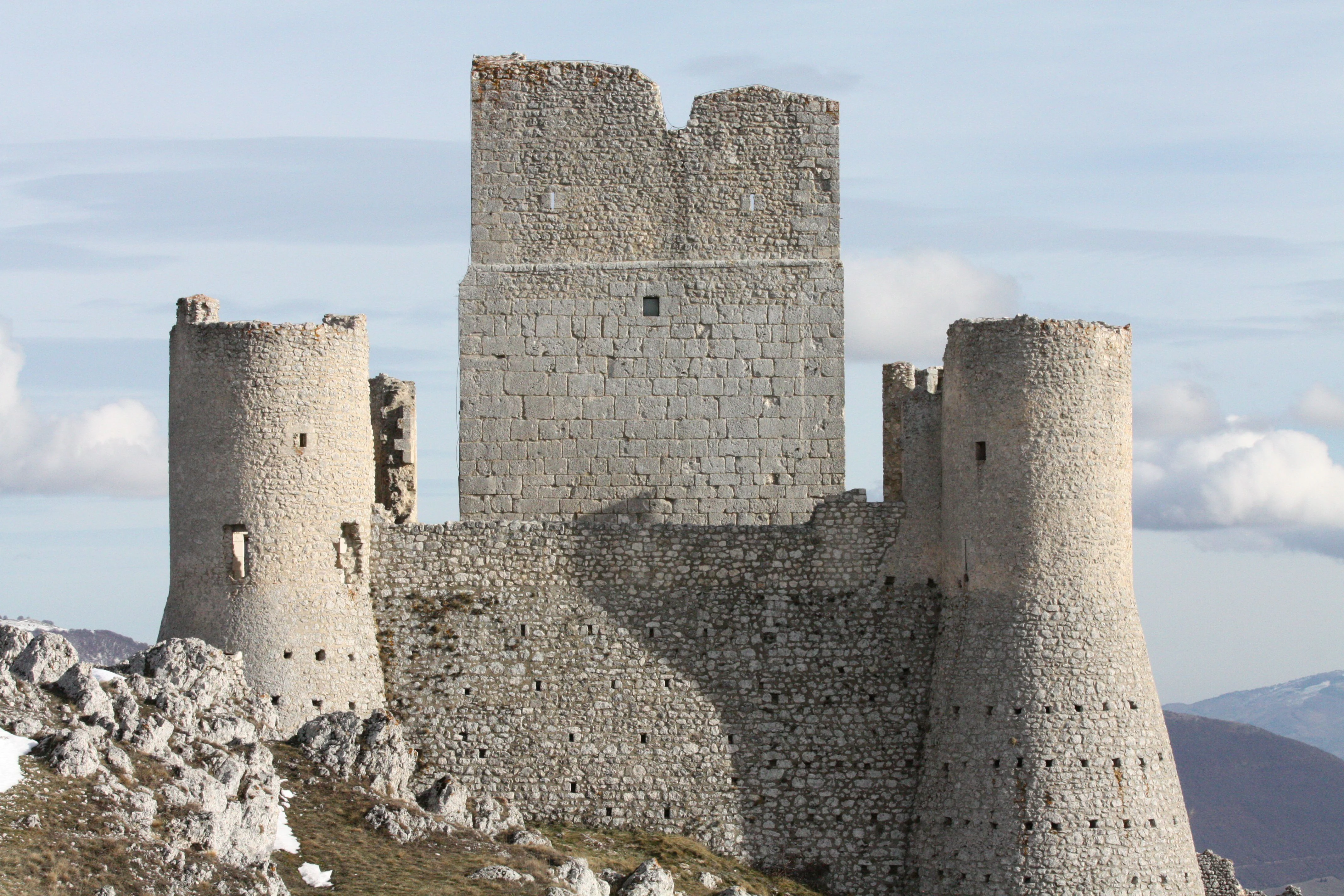 Stone castle ruins with multiple cylindrical towers on a rocky hillside, mountainous background under a partly cloudy sky