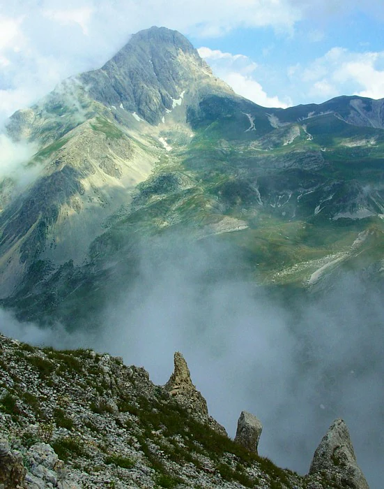 Mountainous landscape with rocky peaks, green slopes, and misty clouds under a partly cloudy sky