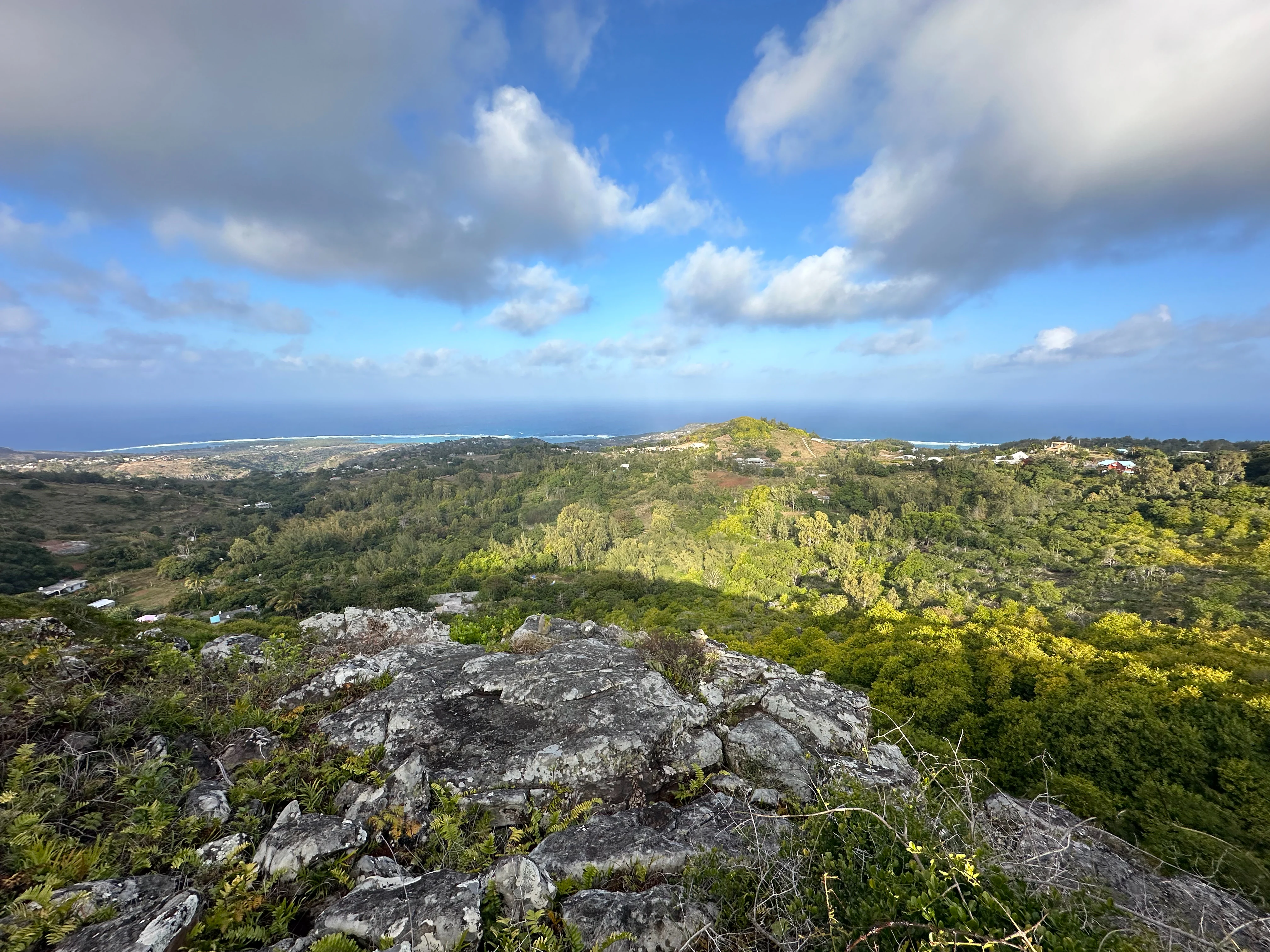 Vista panorámica en ángulo elevado desde una cresta rocosa que contempla colinas verdes y frondosas y bosques hacia el lejano océano azul y la costa de la isla.