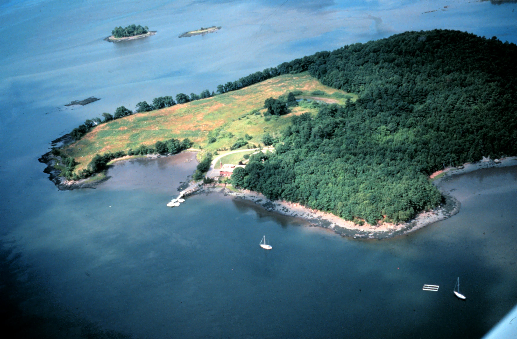 An aerial perspective of a wooded peninsula extending into a large bay, featuring a mix of forest and open grassy areas, with small boats anchored in the surrounding water.