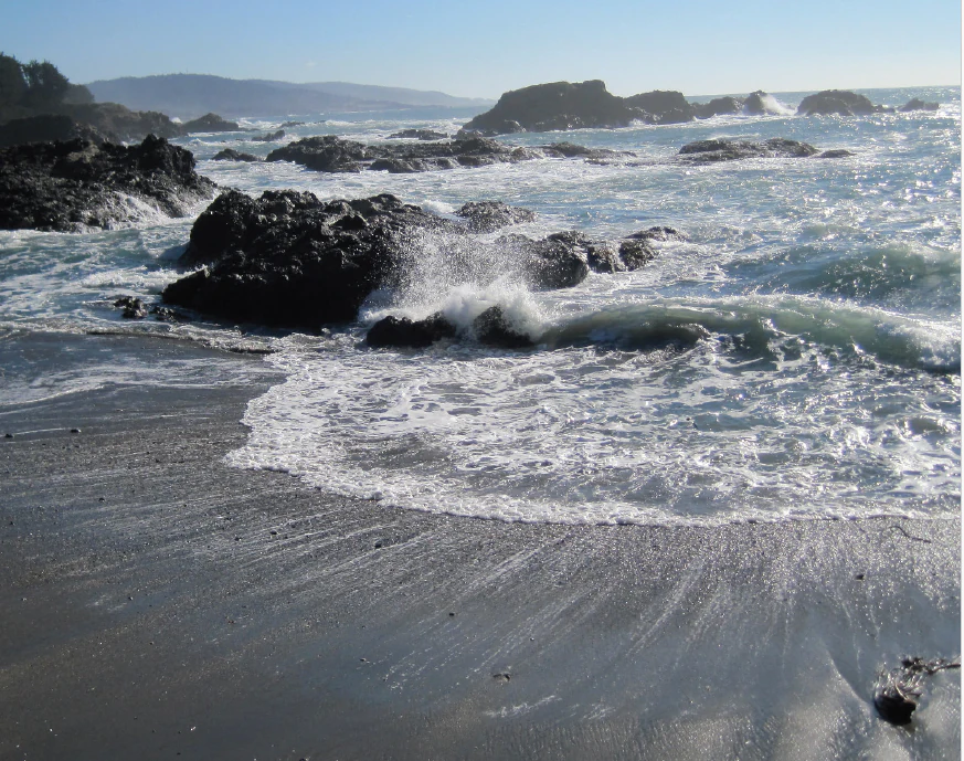 Una escena costera soleada que muestra olas rompiendo sobre rocas oscuras y afiladas en una playa de arena, con colinas lejanas visibles a lo largo del horizonte.