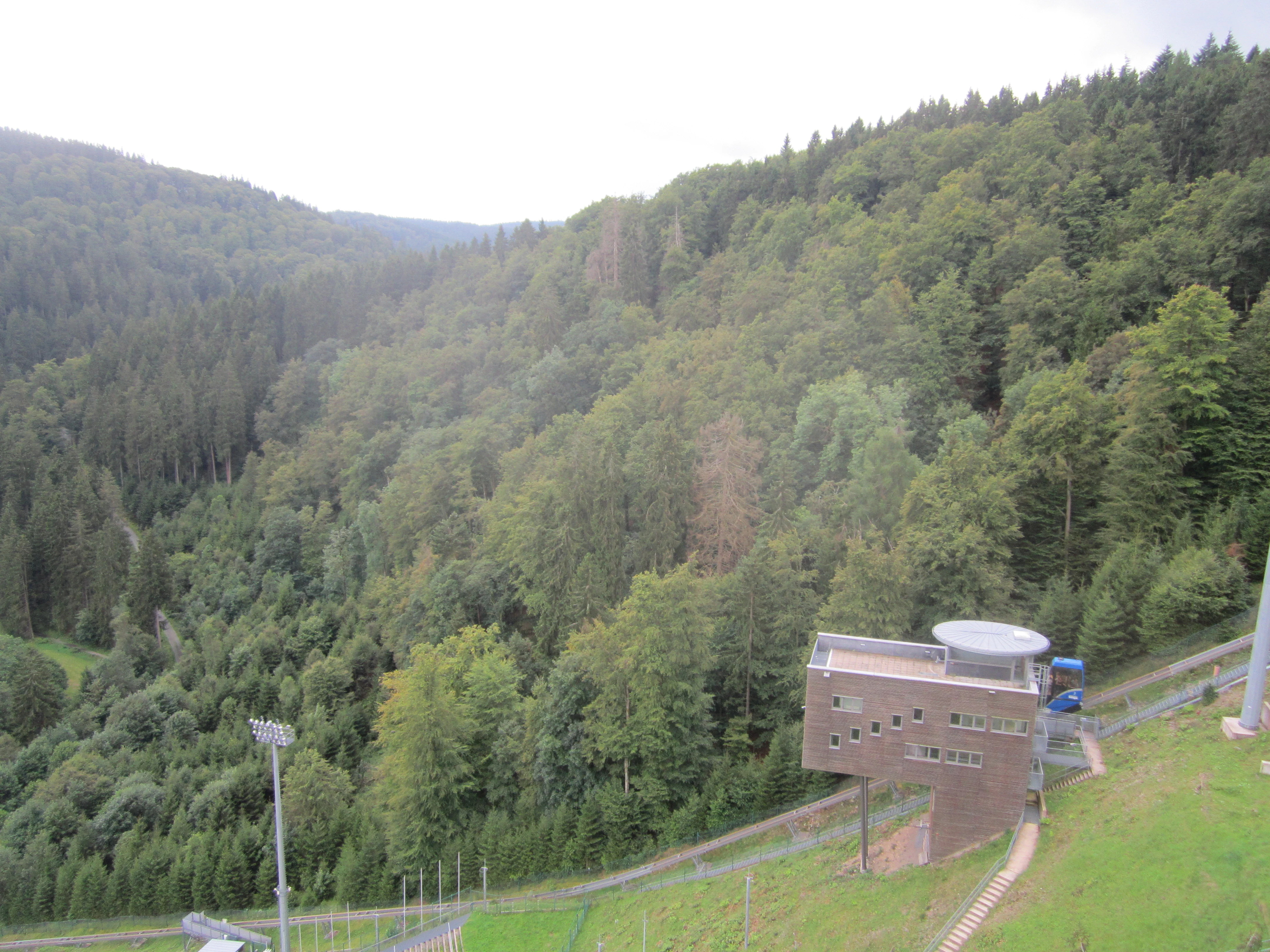 A wide shot showing a densely forested hillside rising in the background with a modern brick judge tower building in the lower right foreground.