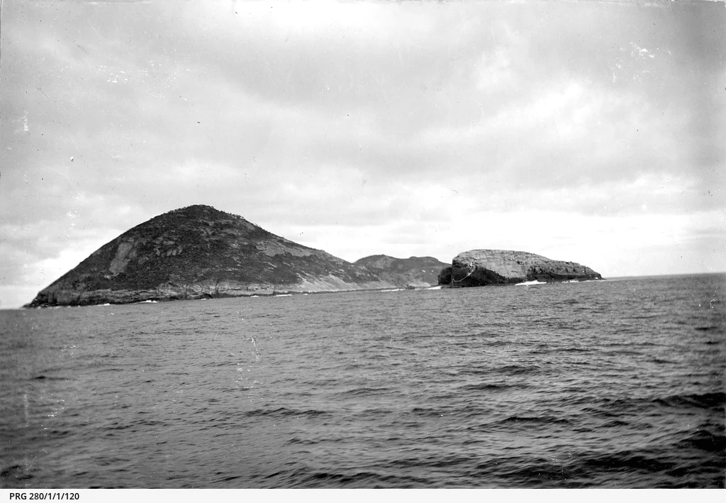 A black and white photograph showing a distant view of Greenly Island, a rugged island with a prominent peak surrounded by the open ocean under a cloudy sky.