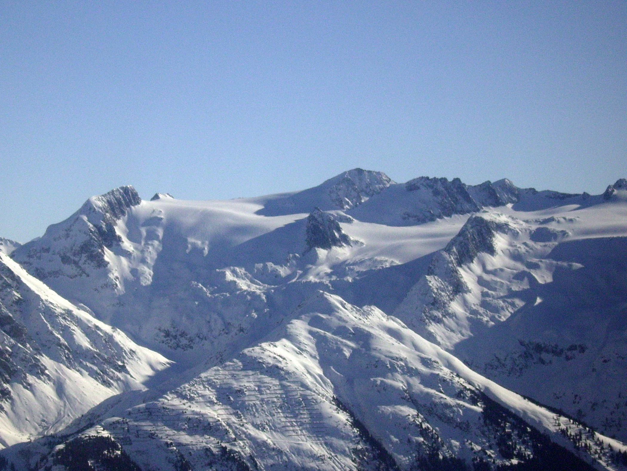 A wide mountain landscape showing the snow-covered peak of Piz Medel and the adjacent glacier slopes against a clear blue sky.