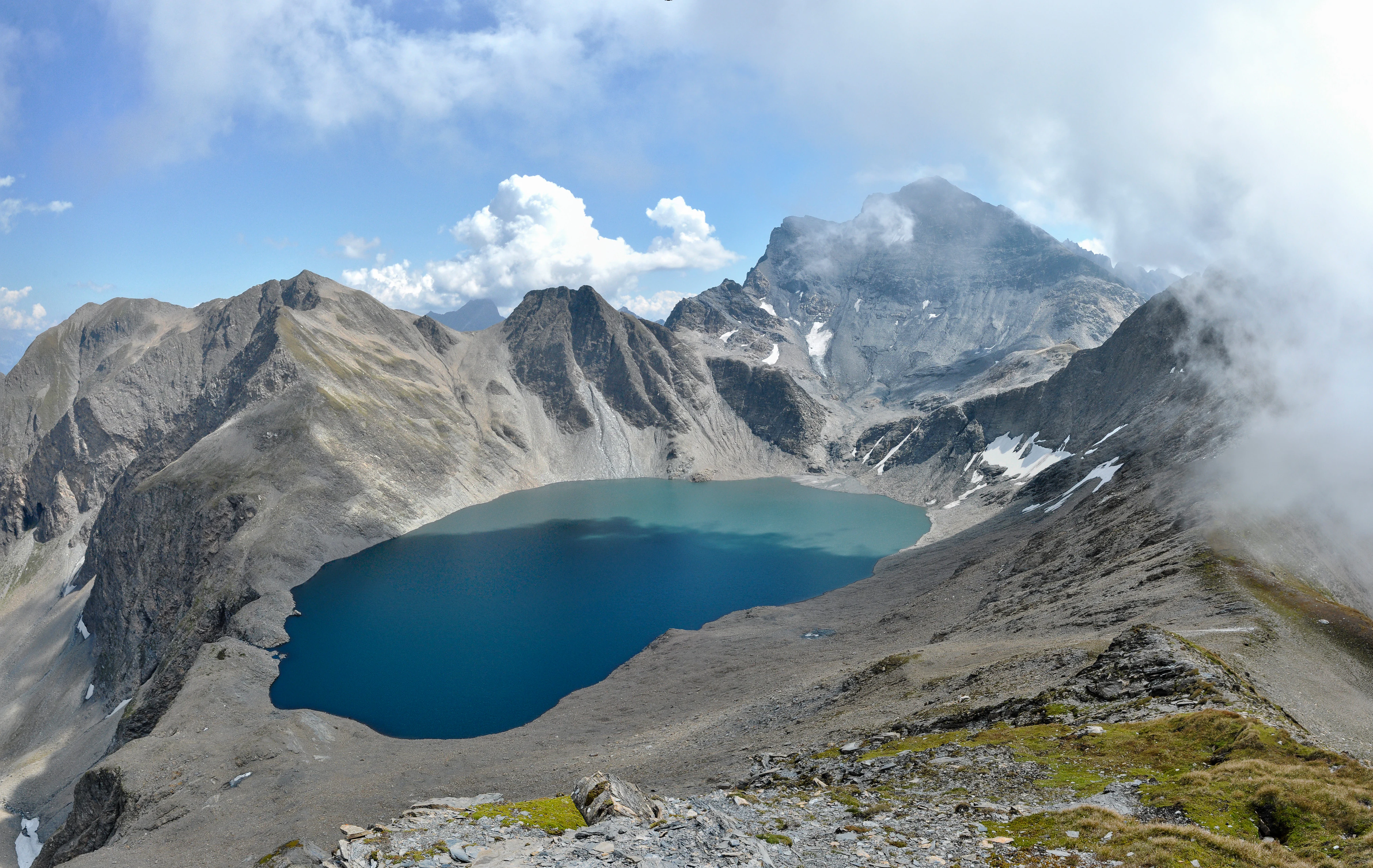 A wide, high-angle view of an alpine glacial lake with dark blue water, surrounded by rocky peaks and ridgelines under a partially cloudy sky.