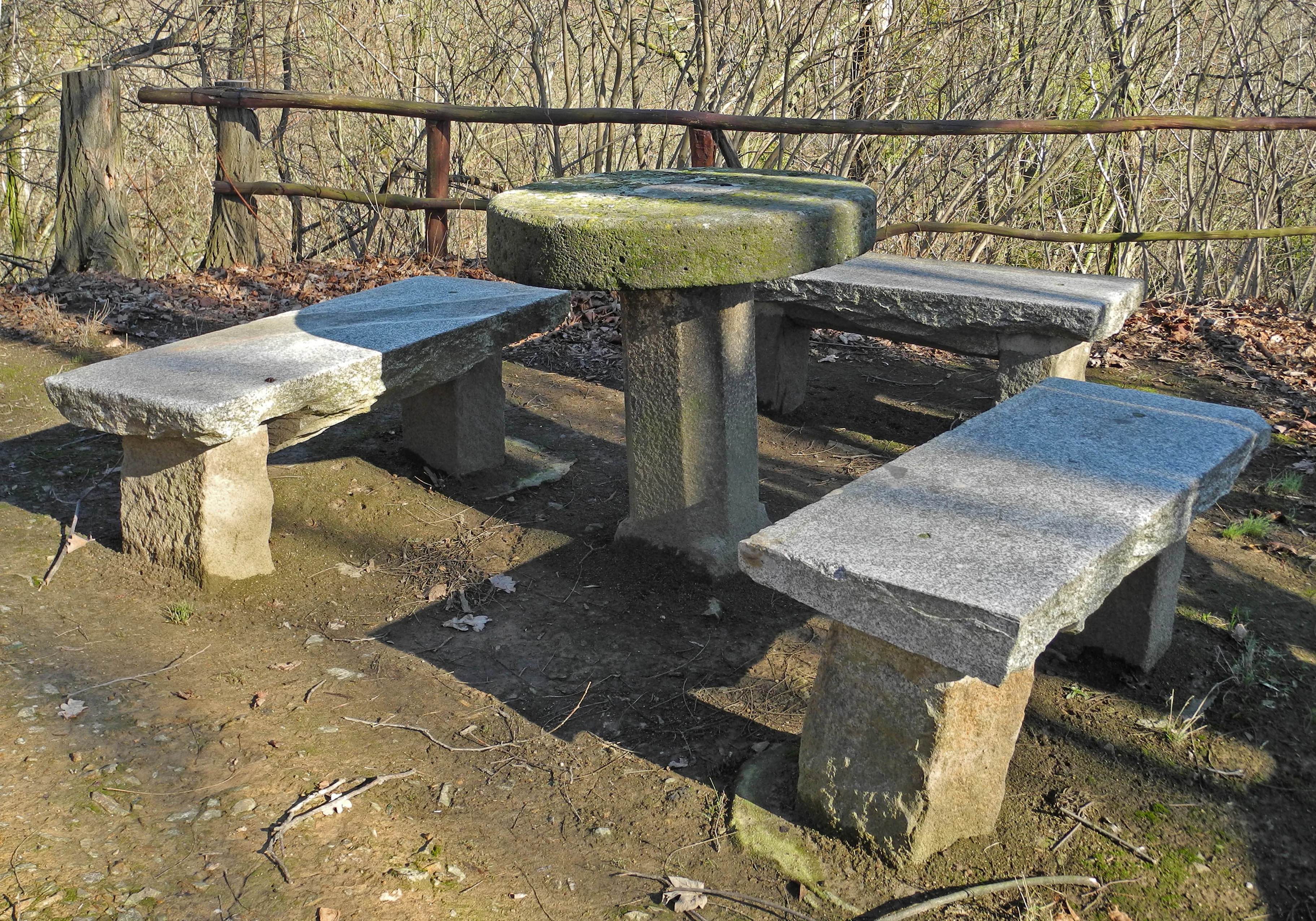 A rustic outdoor seating area featuring a round stone table surrounded by matching stone benches, located on a dirt path with a simple wooden railing in the background.