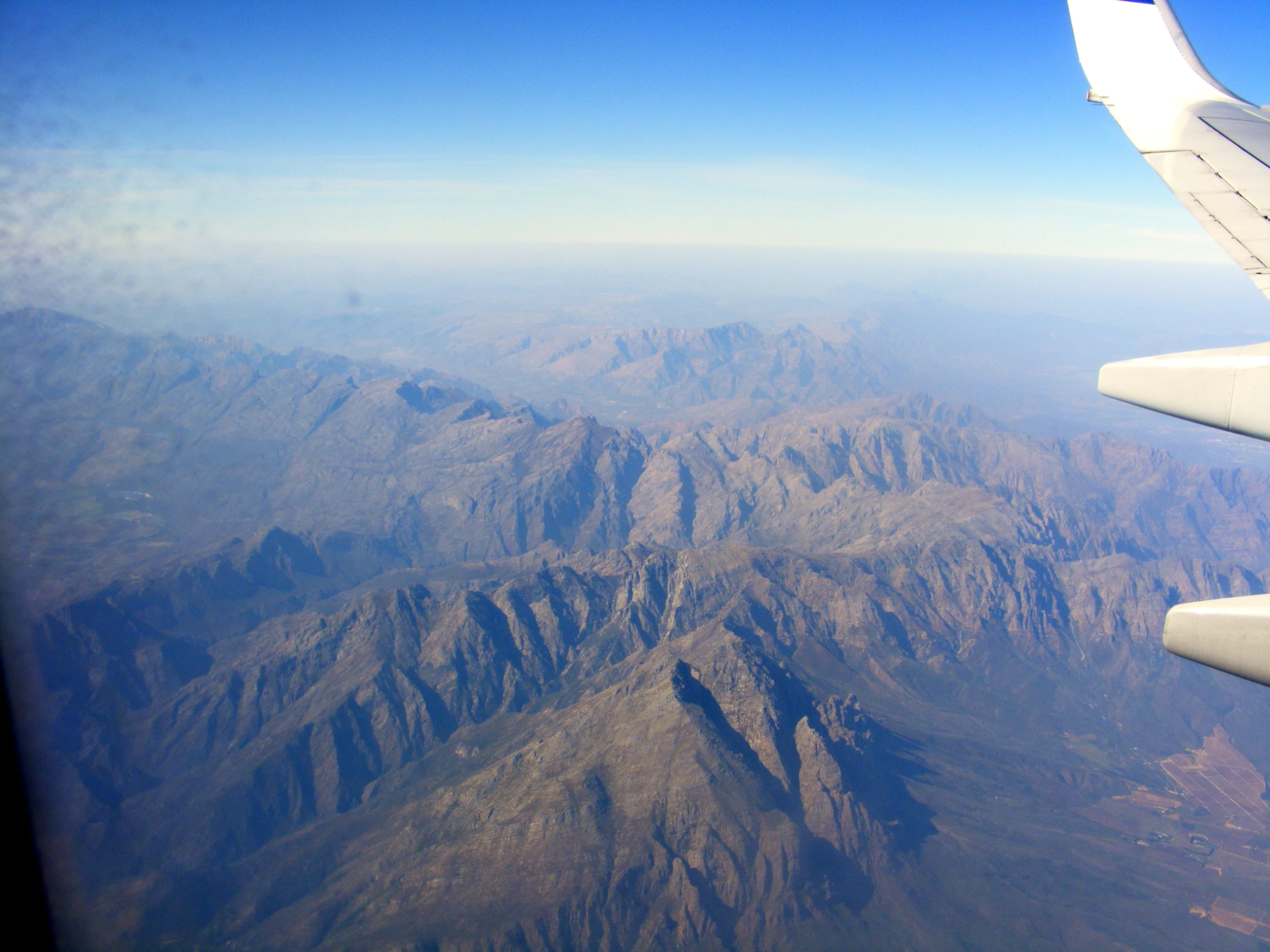 A high-altitude aerial view of a rugged, dry mountain range with deep valleys and ridges, taken from an airplane window with the wing visible on the right side.